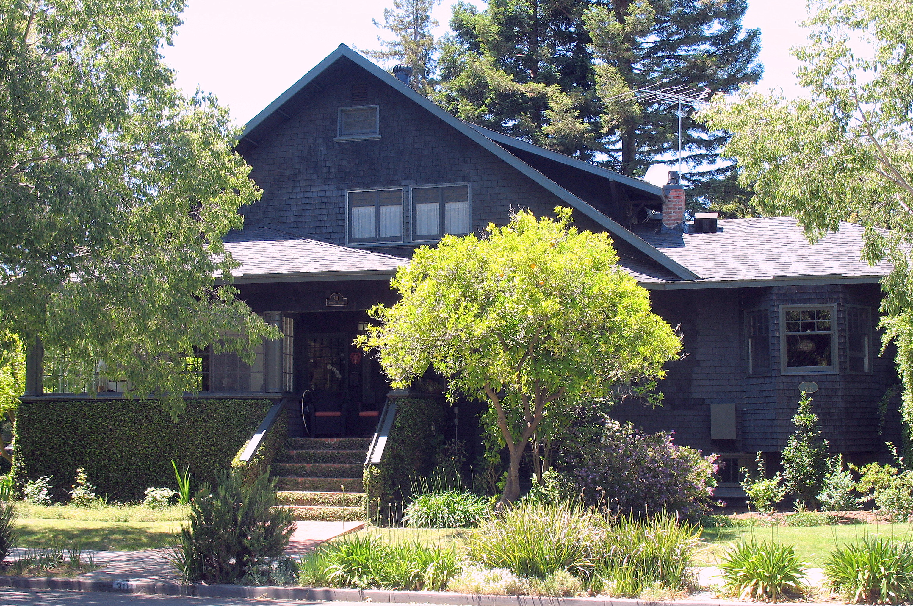 A dark-shingled Craftsman house with a covered porch surrounded by mature trees and landscaping.