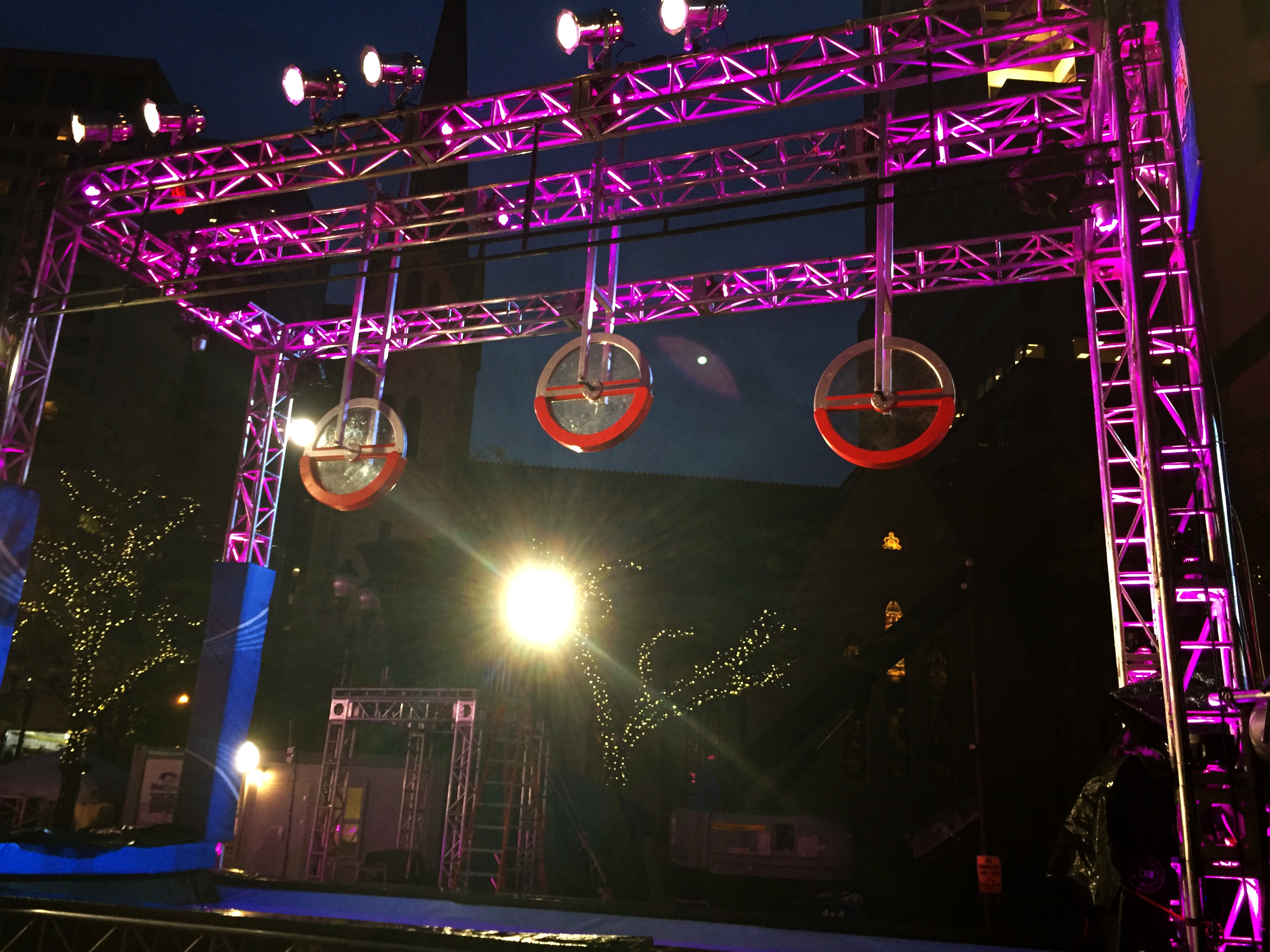 Three wheel-shaped obstacles hang from a pink-lit metal truss on a Ninja Warrior course at night.