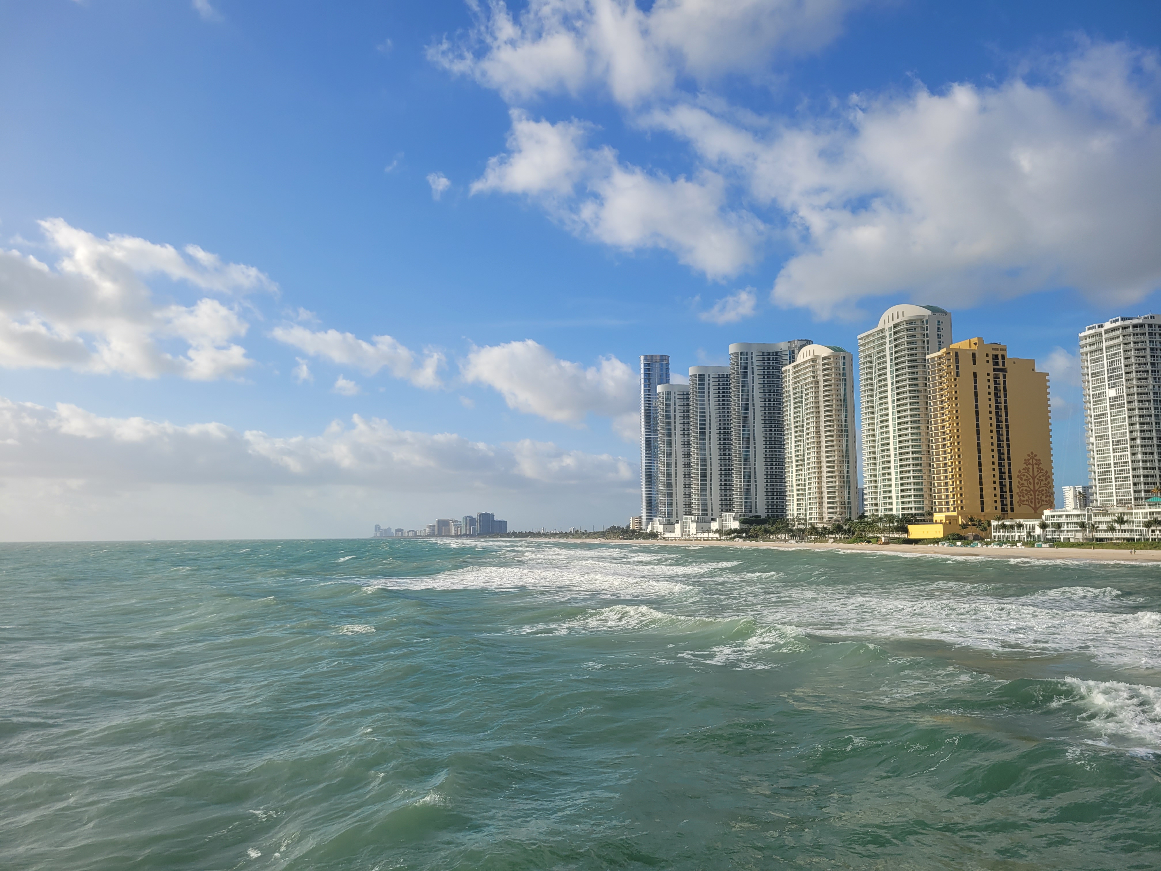 High-rise towers line a South Florida beachfront with turquoise ocean waves in the foreground.