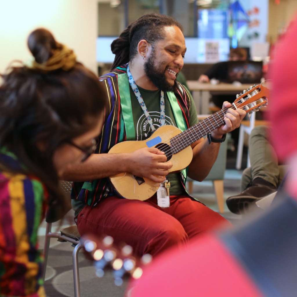 A smiling man with dreadlocks plays a small guitar in an indoor community space.