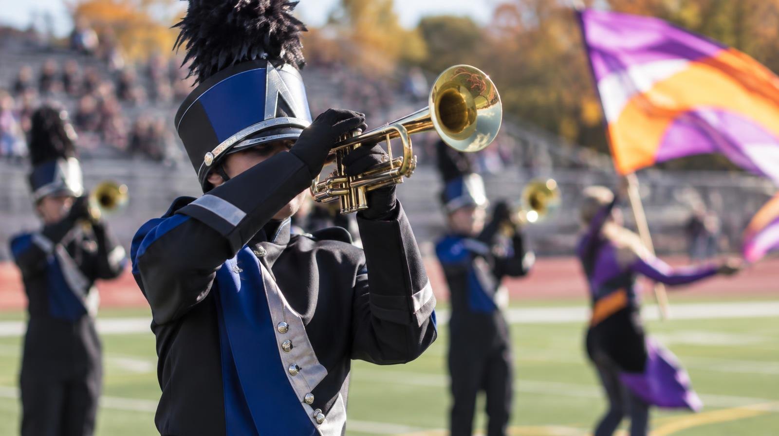 Marching band in colorful uniforms performing on a football field with color guard flags.