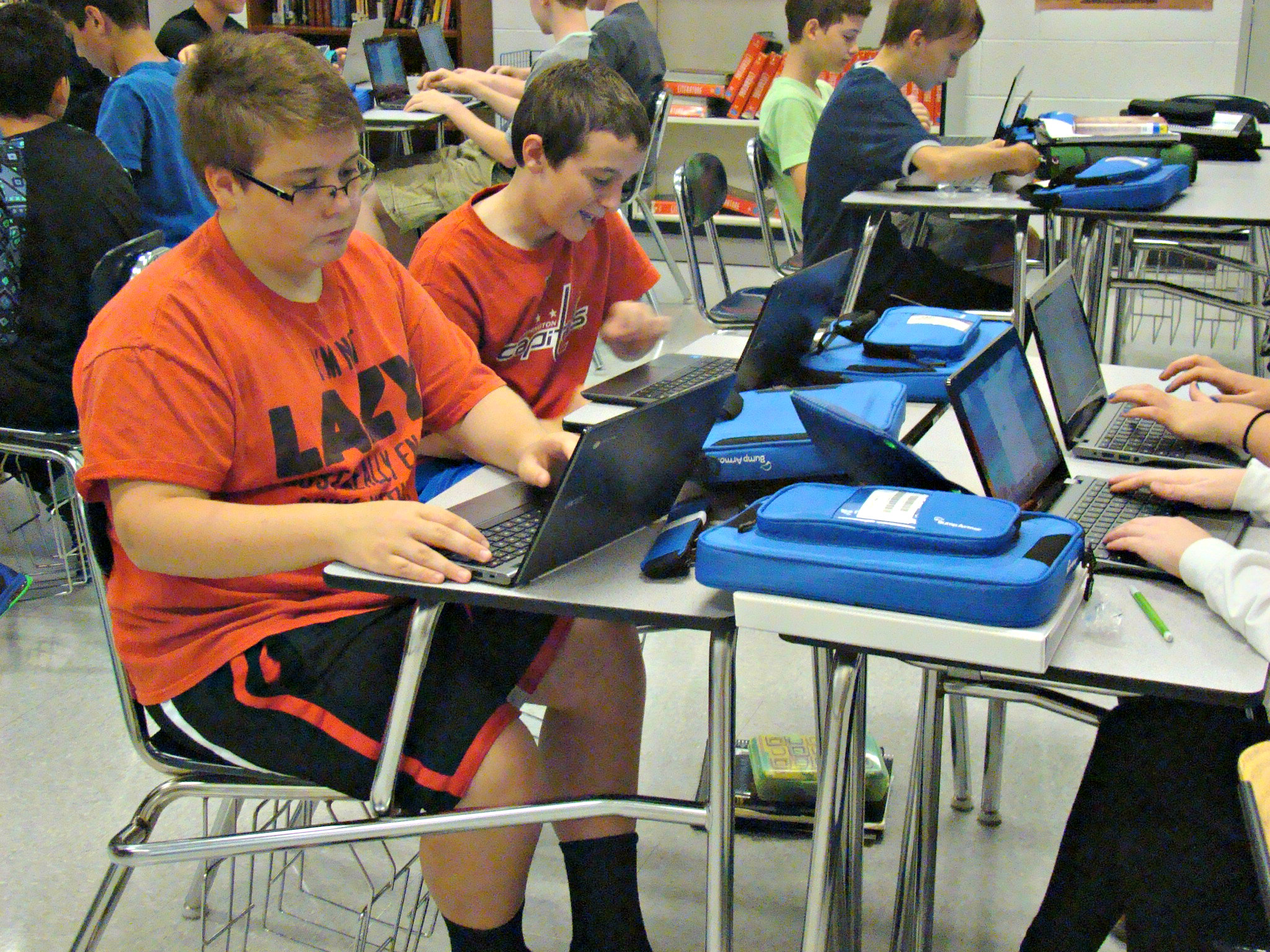 Two students in orange shirts use Chromebooks at school desks in a classroom.
