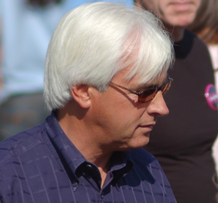 White-haired man in sunglasses and blue shirt at a racetrack.