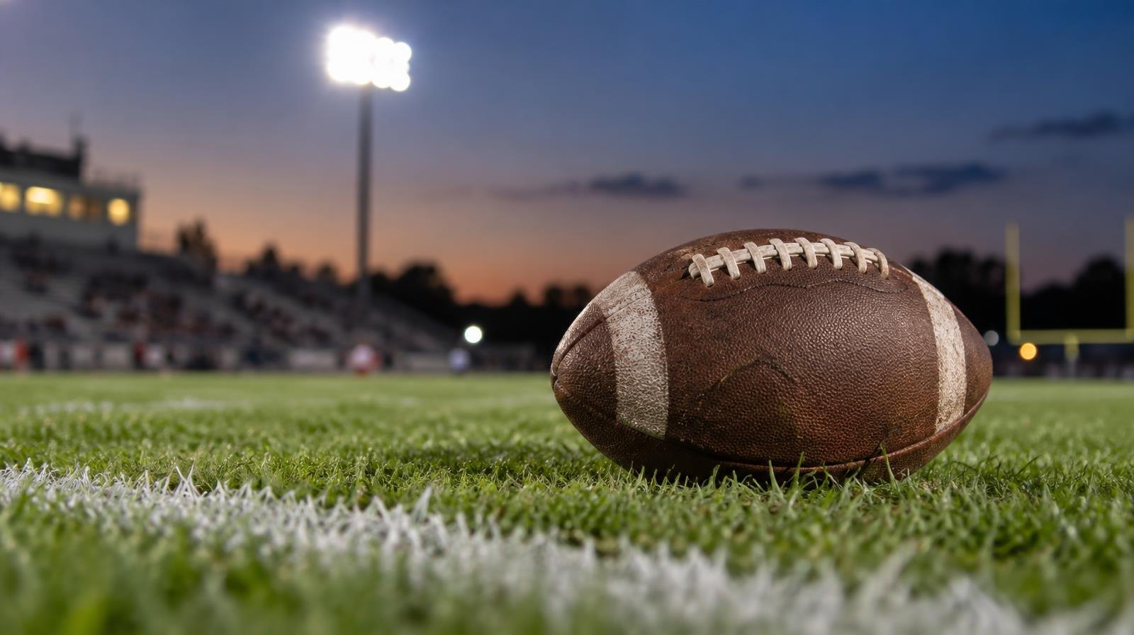 An American football resting on green grass near a white yard line under stadium lights.