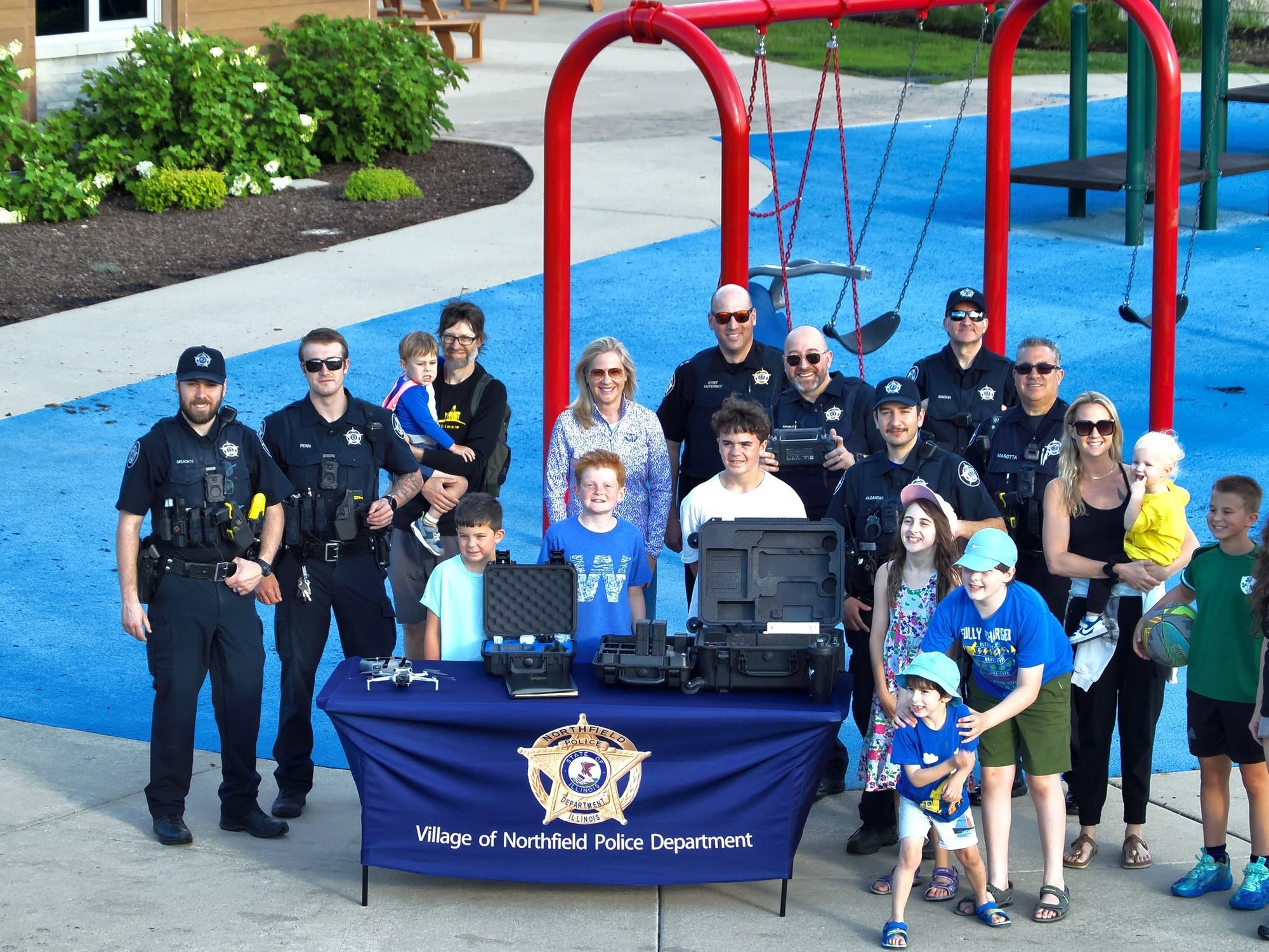 Police officers and community members, including children, gathered around a table with police equipment at a playground.