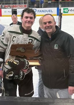 Two people pose on an ice rink, one holding a wooden championship plaque.