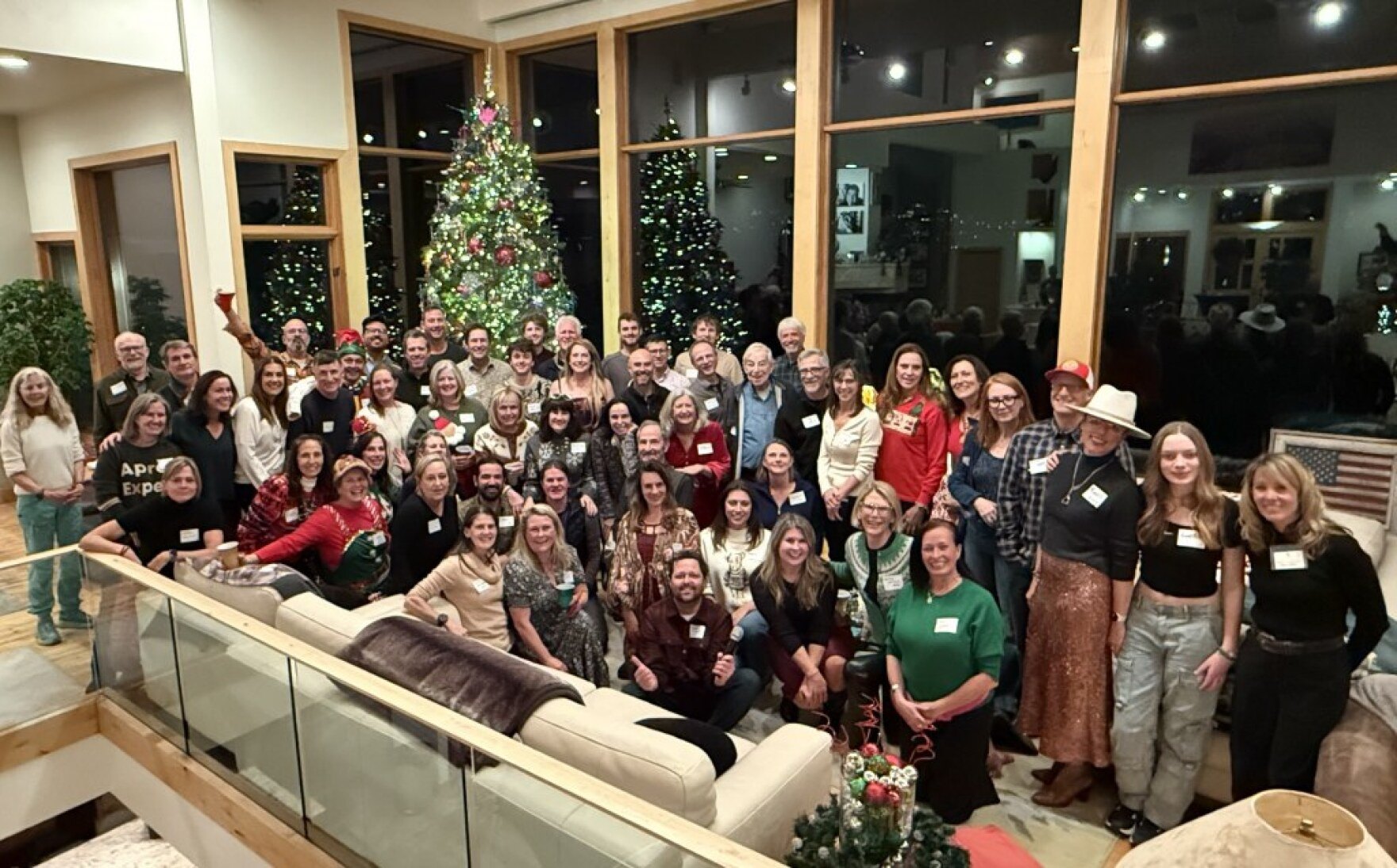 Large group of adults posing indoors in front of a decorated Christmas tree, many wearing name tags.