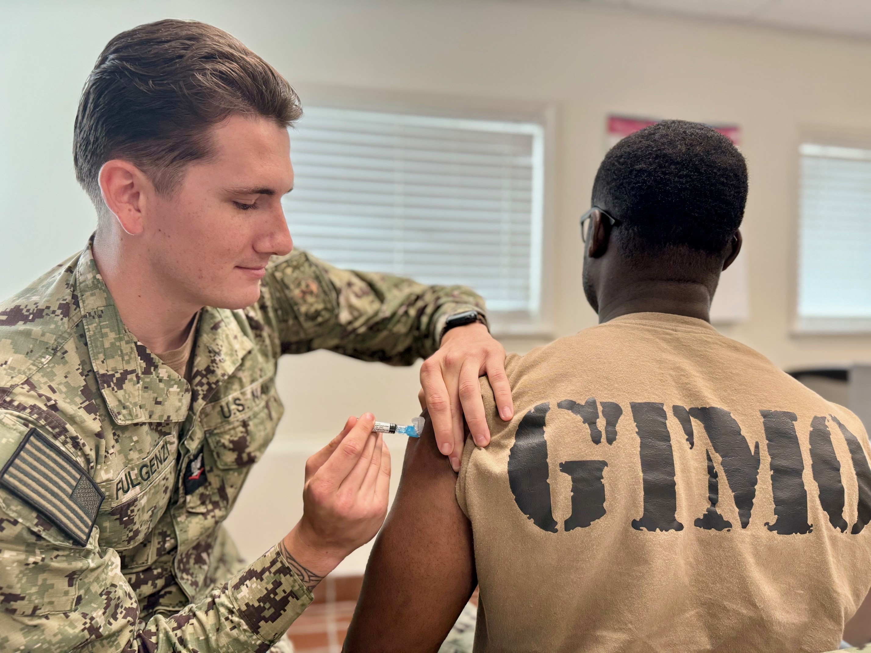 A healthcare worker in uniform gives a flu shot to a person's upper arm indoors.
