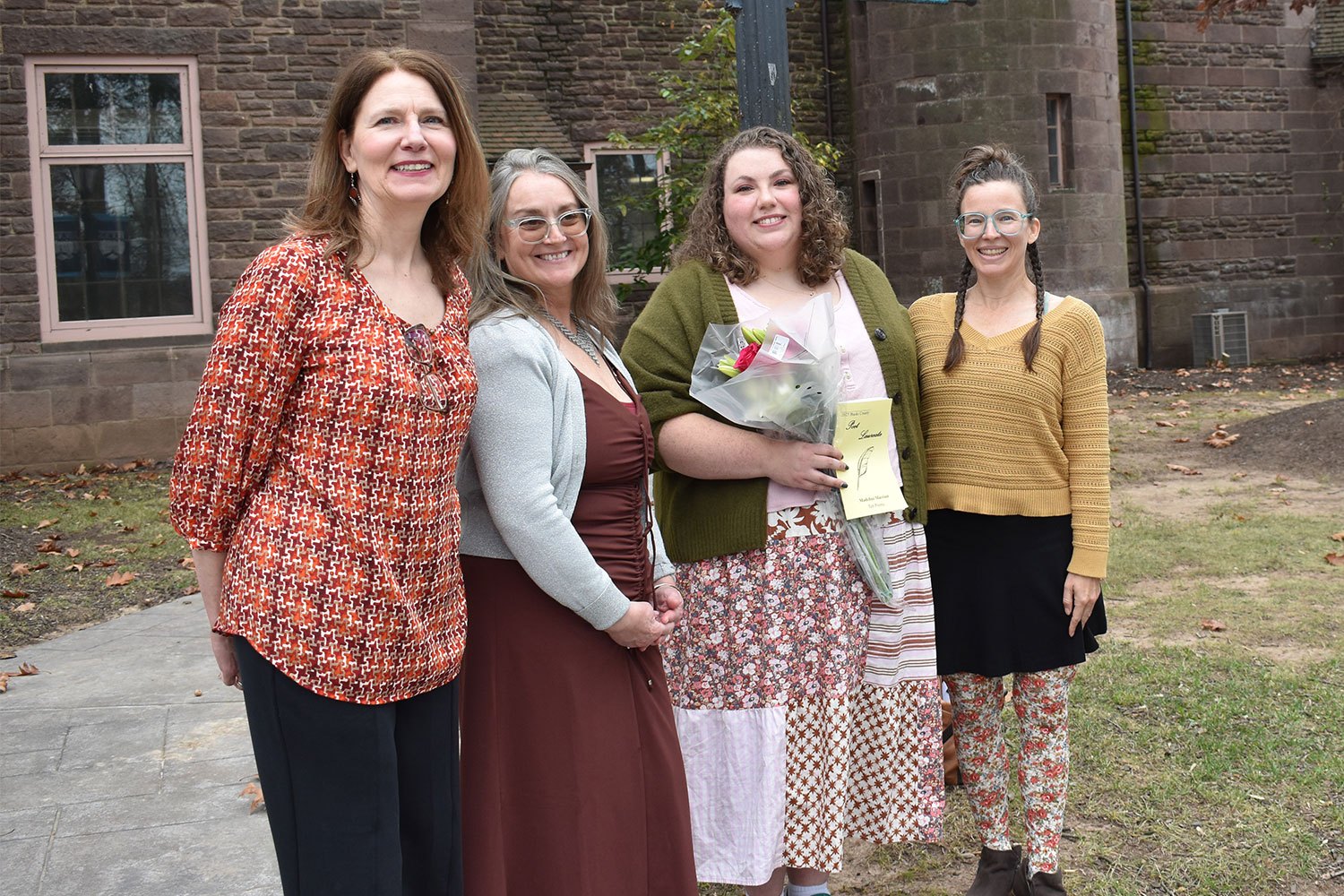 Four women pose together outside a historic stone building, one holding a bouquet of flowers.
