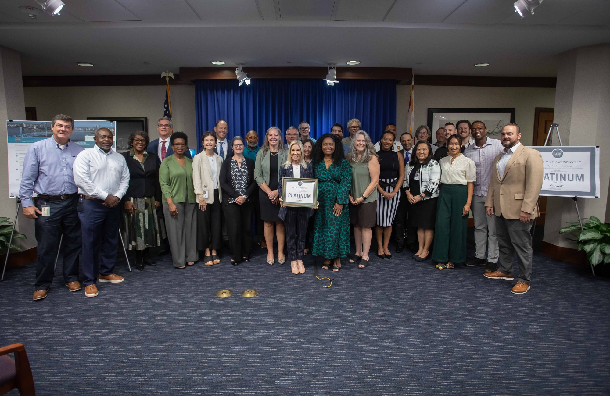 Large group of city employees posing indoors with a framed certificate and a Platinum sign on an easel.