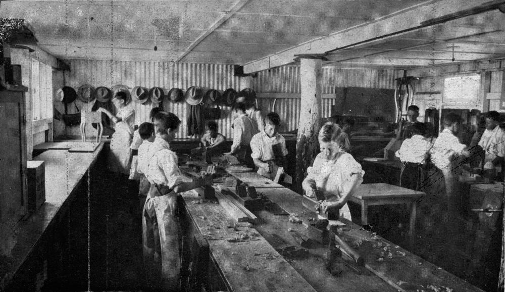 Students in aprons work at wooden benches with hand tools in a classroom.