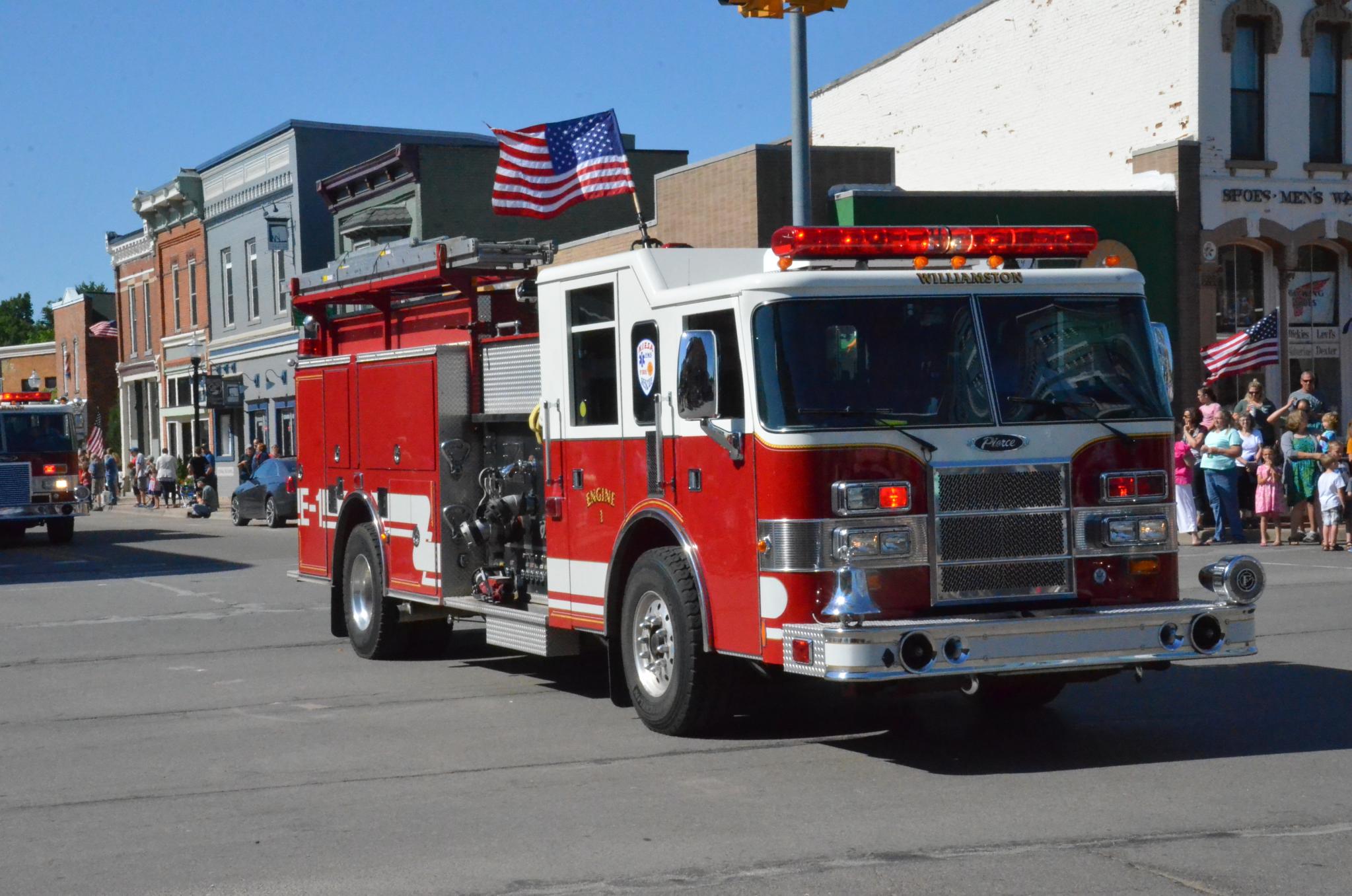 Red and white fire engine with an American flag mounted on top driving down a street.