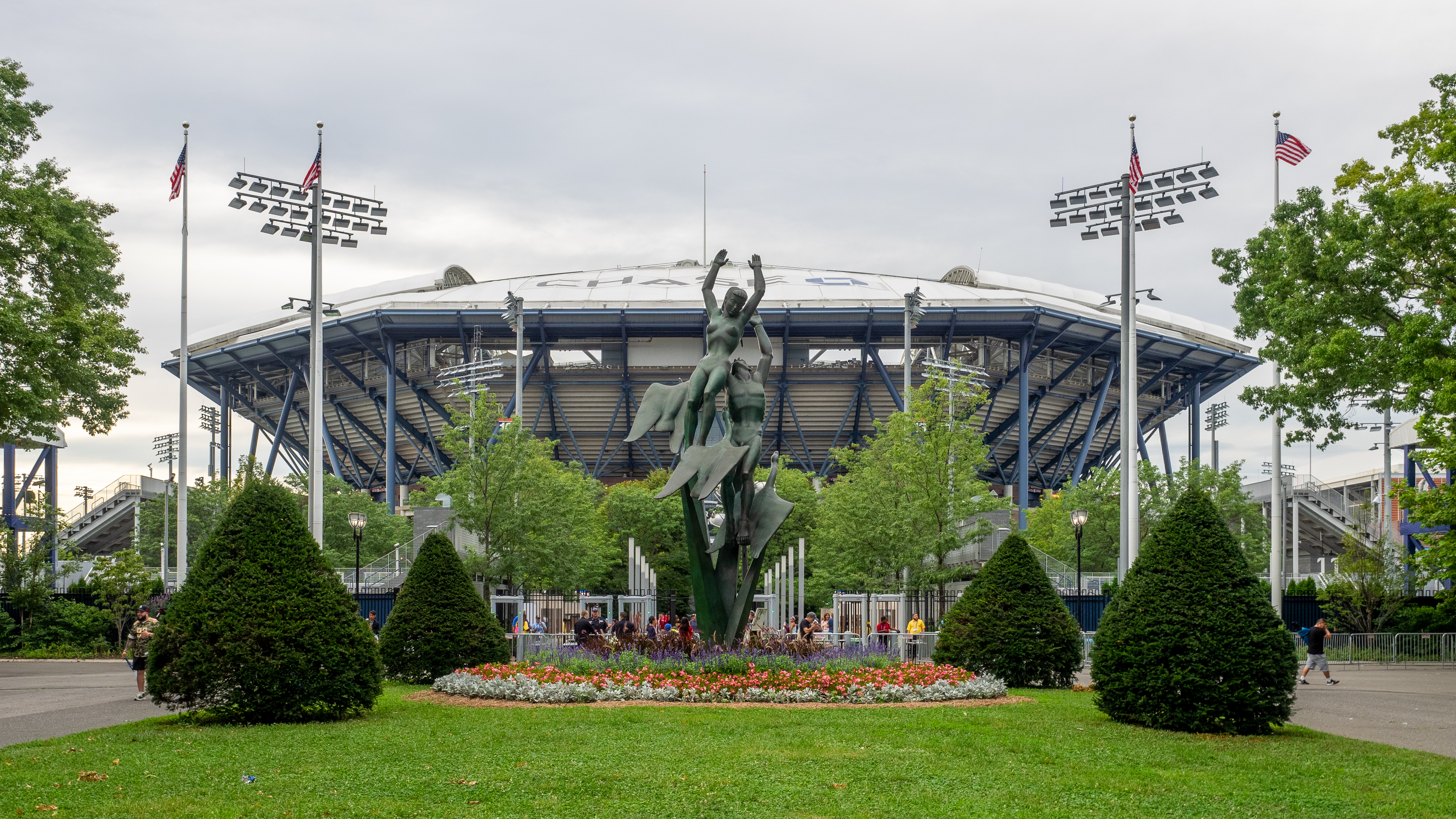 Exterior of a large domed tennis stadium with a bronze sculpture and American flags in front.