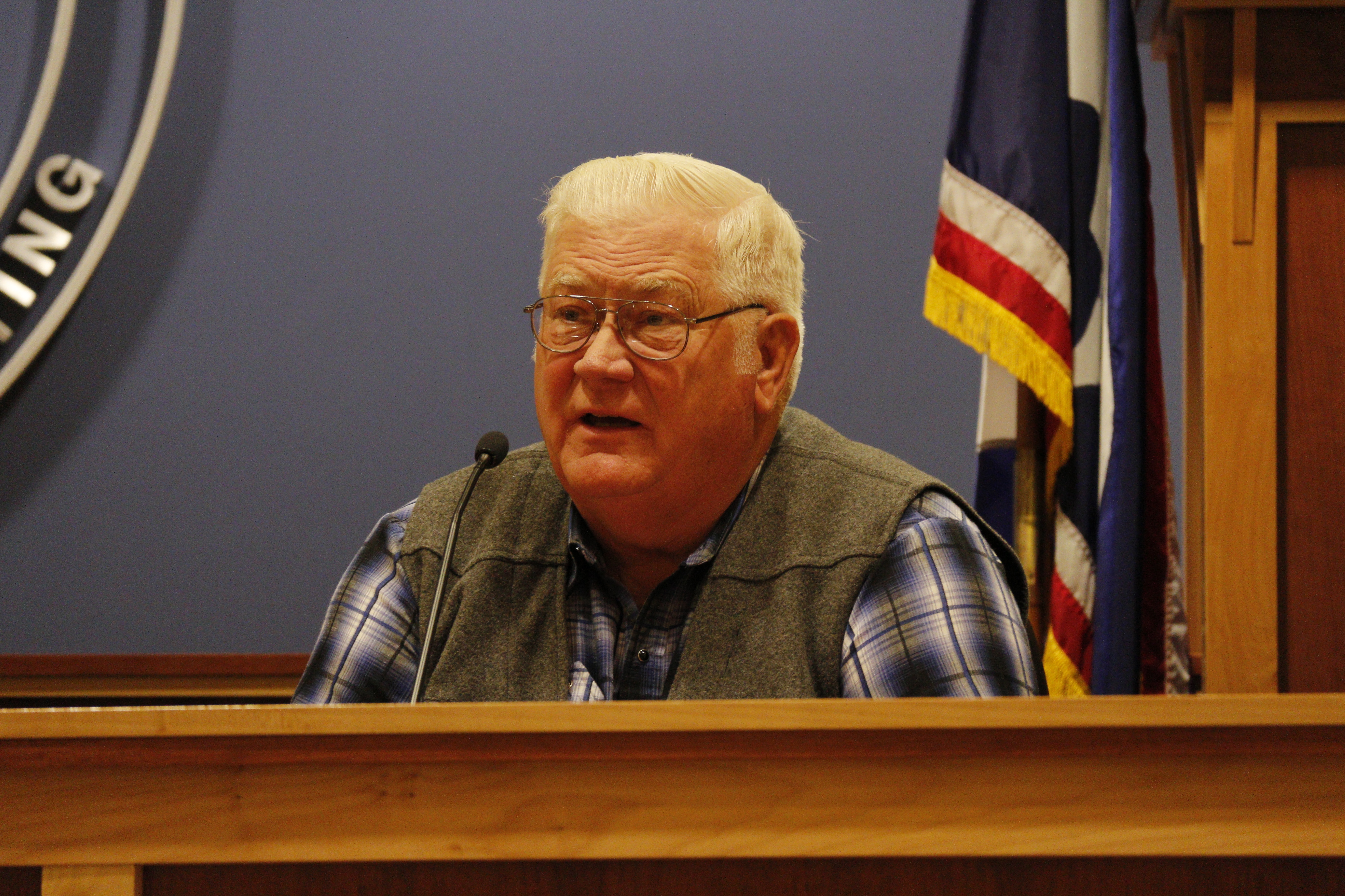 Older man in glasses and plaid shirt speaking at a podium with an American flag behind him.