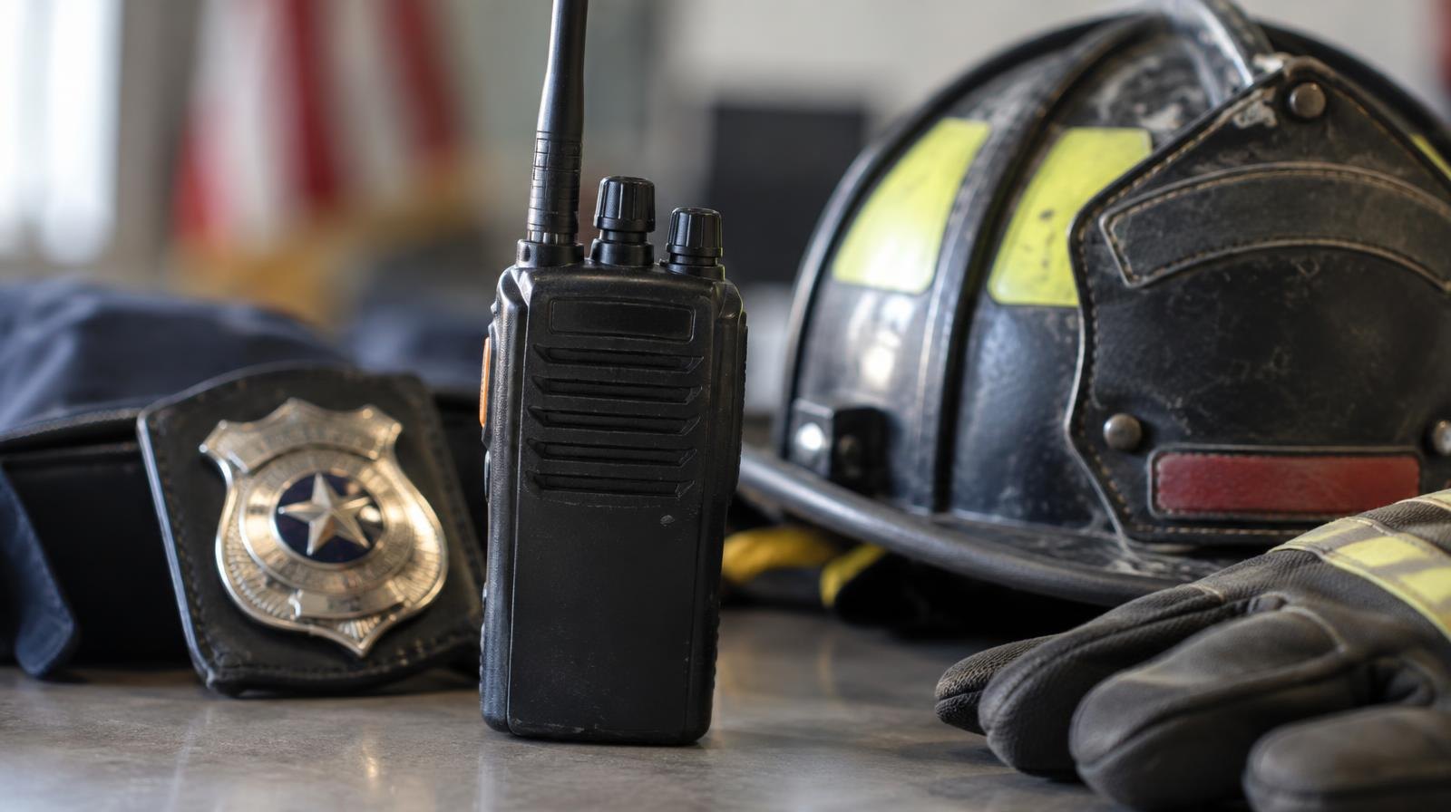 Handheld two-way radio transceiver resting beside a badge and a firefighter helmet.