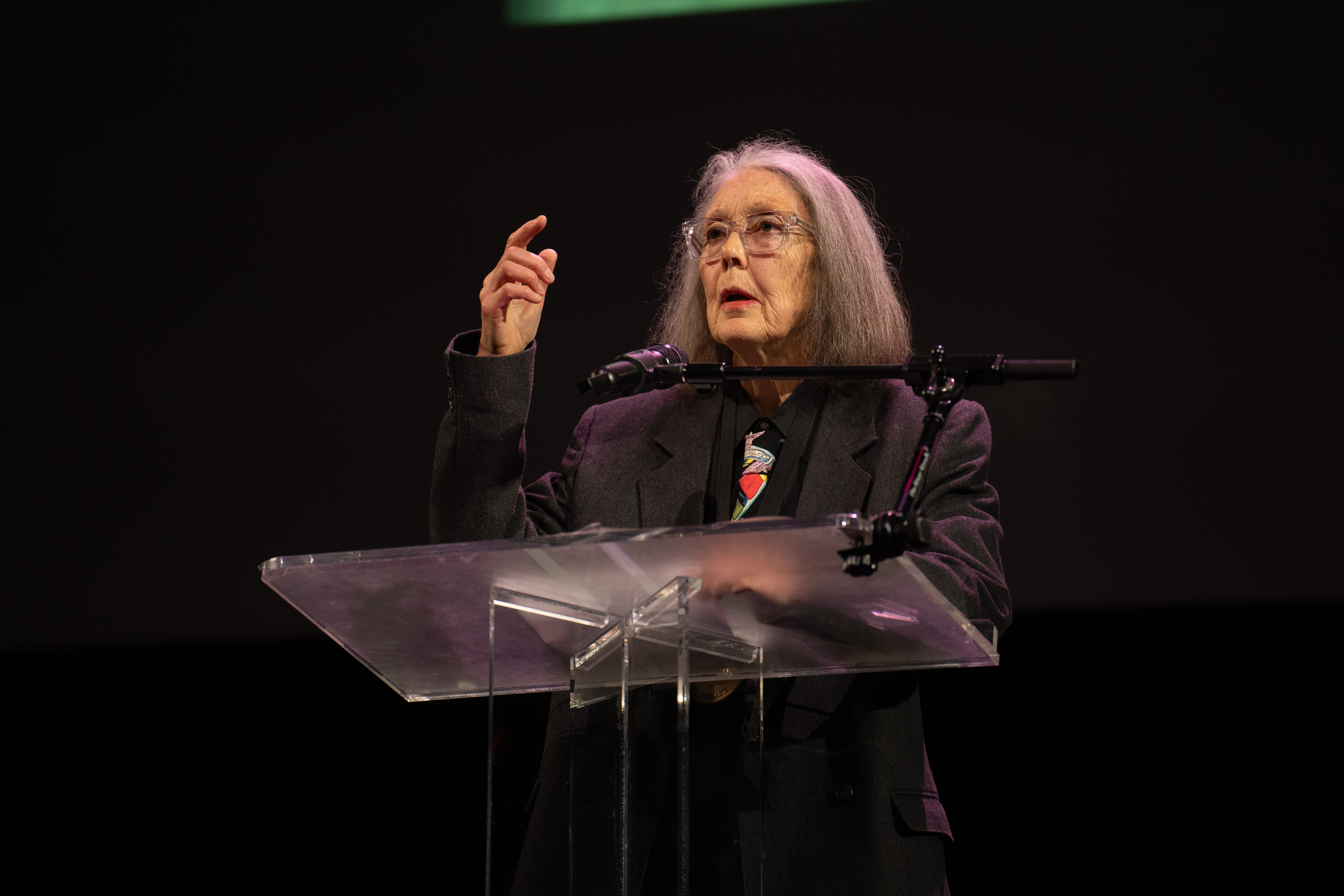 A woman with long gray hair and glasses gestures at a clear podium with a microphone.