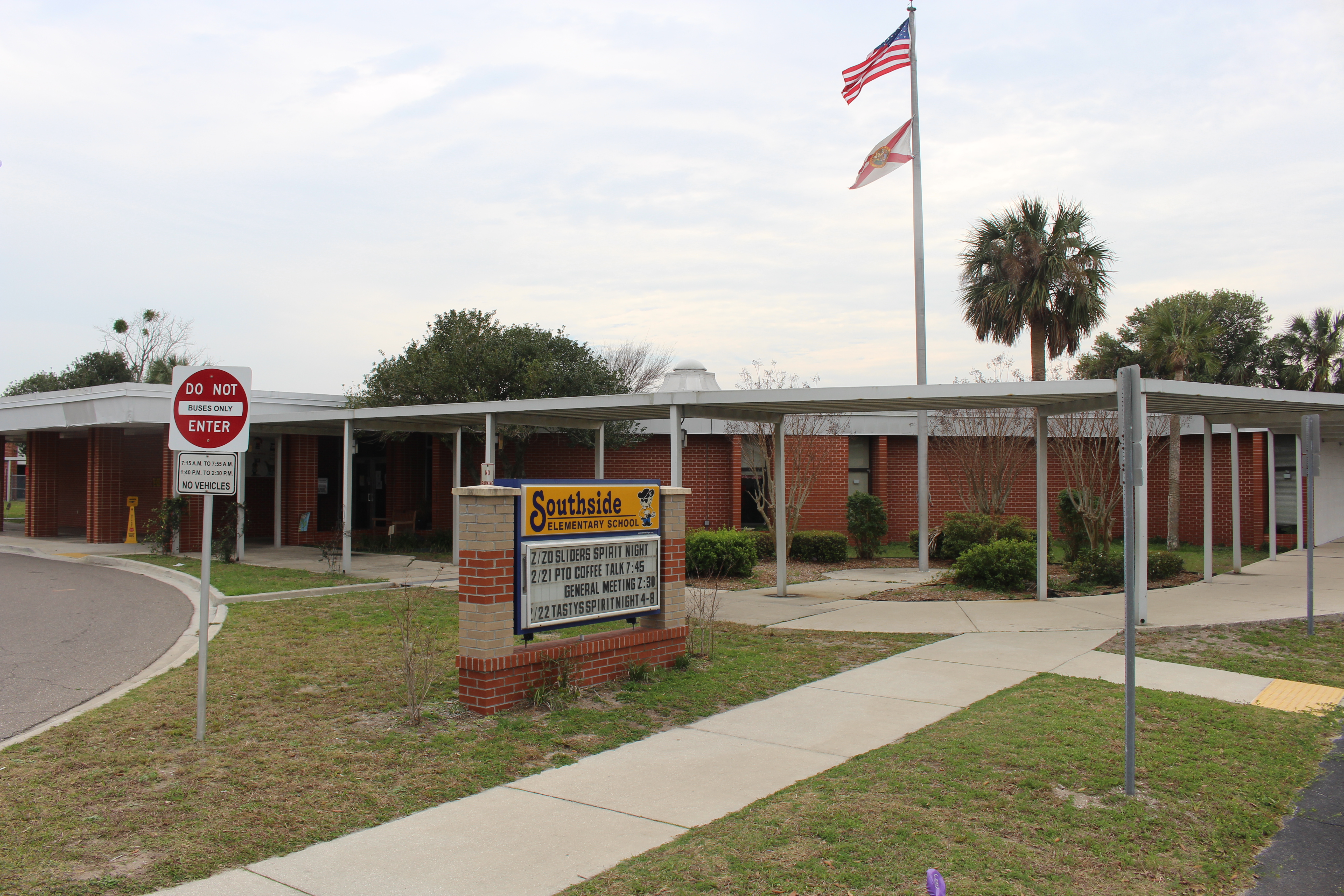 Single-story brick school building with a covered walkway, marquee sign, and flagpole flying US and Florida flags.