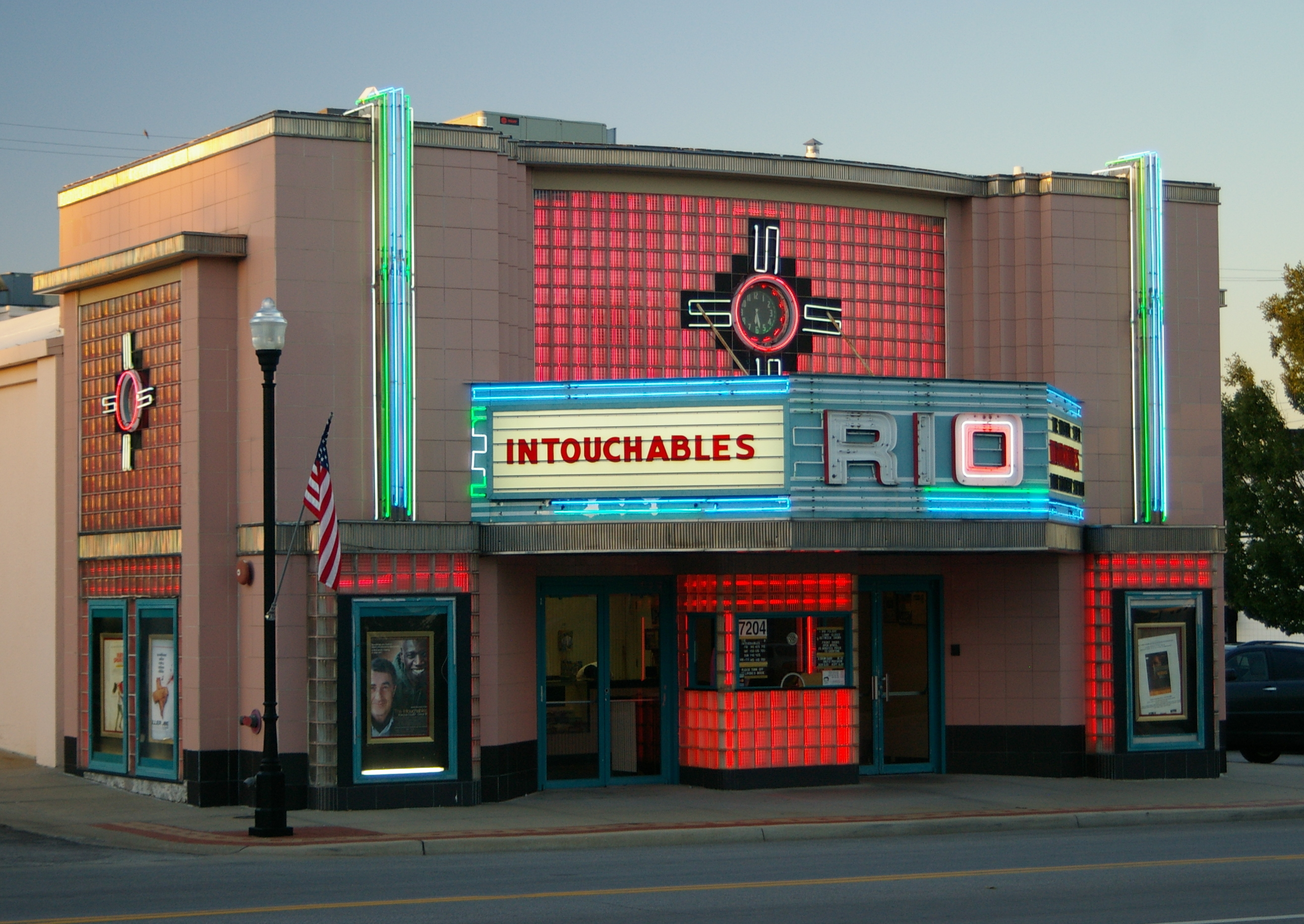 Art Deco cinema facade with teal and red neon signage and a marquee on a city street.