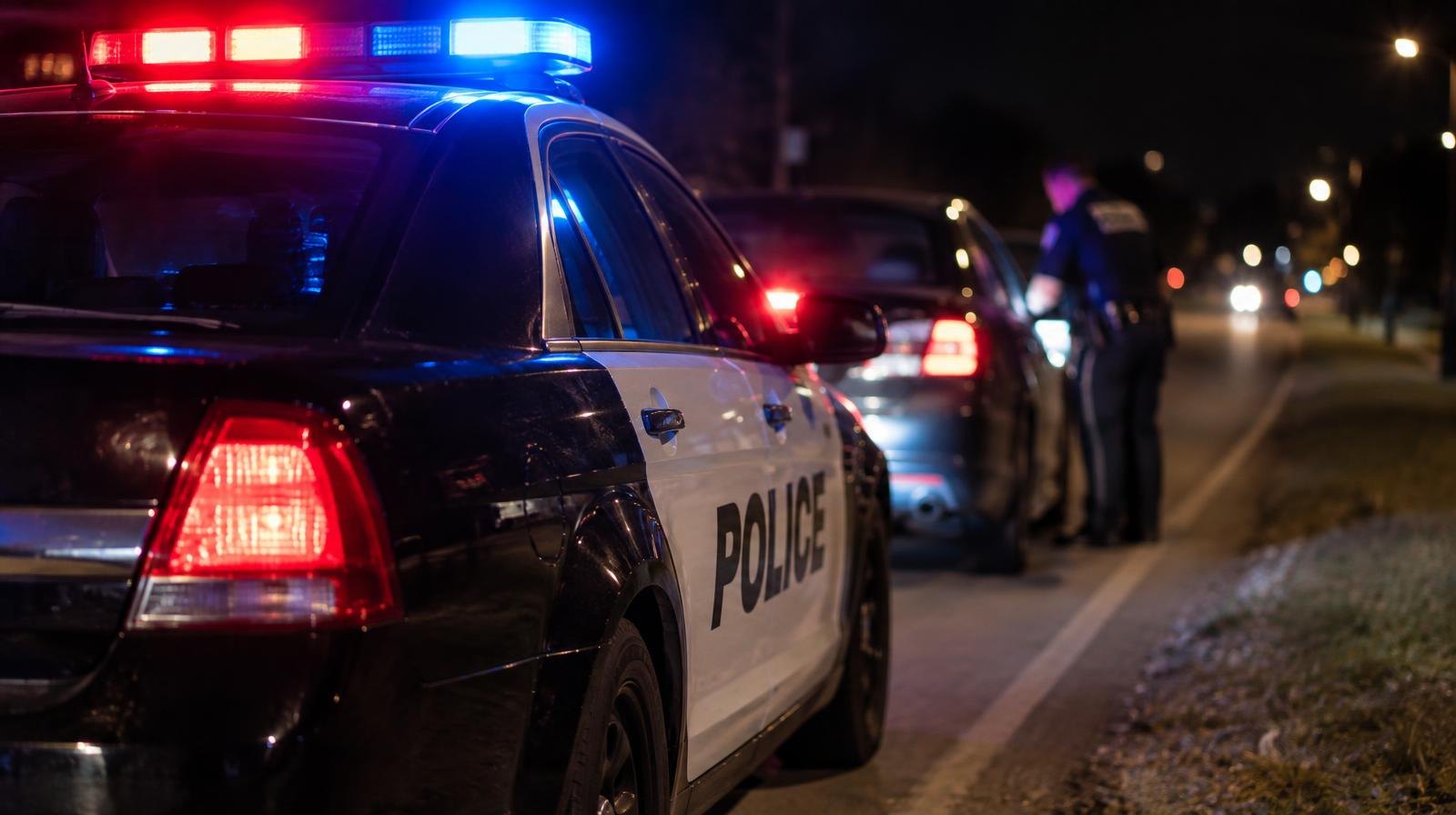 A police car with red and blue emergency lights on, parked on a dark suburban road shoulder.