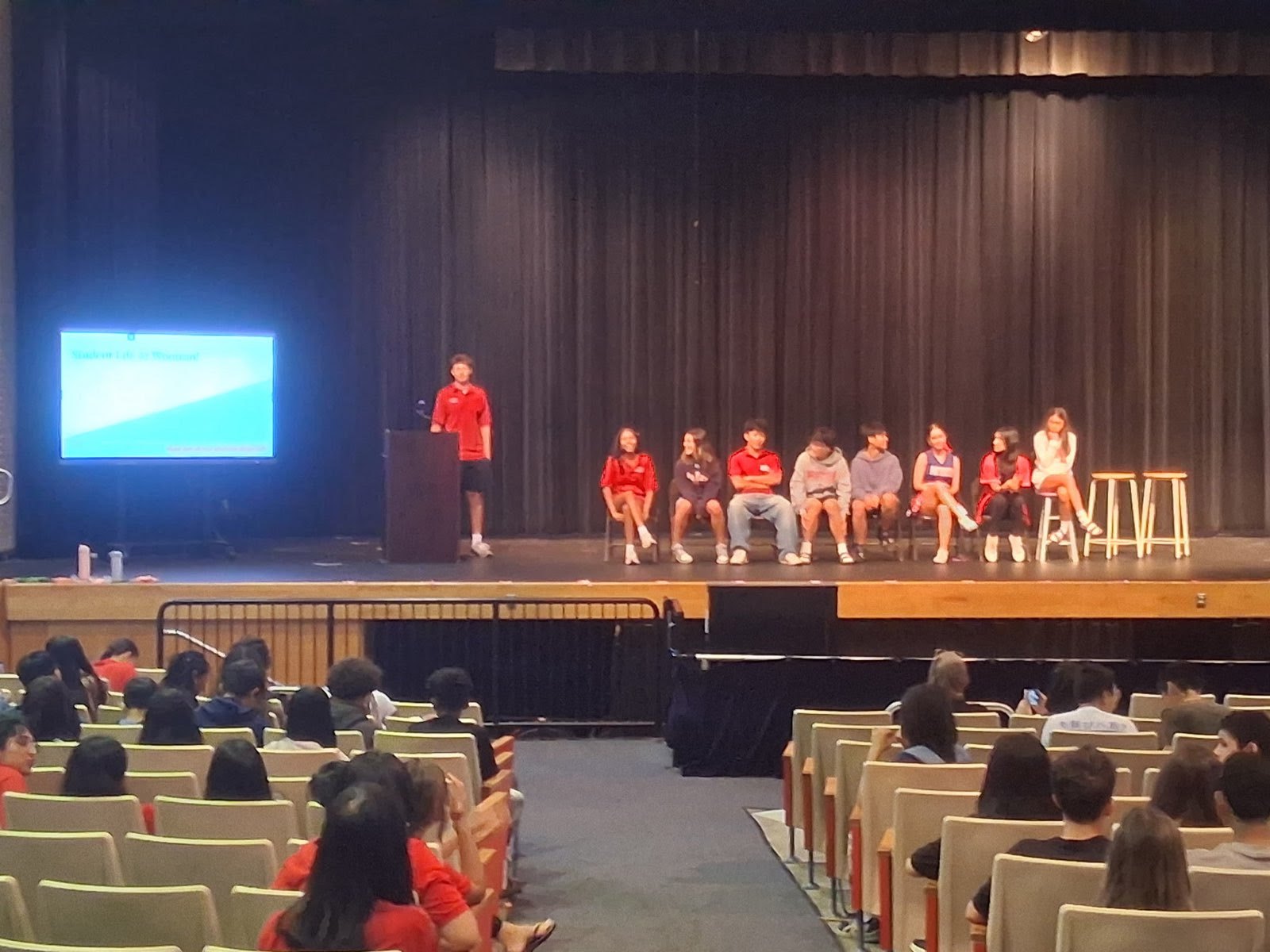A student speaks at a podium on a school auditorium stage while rows of students watch from the audience.