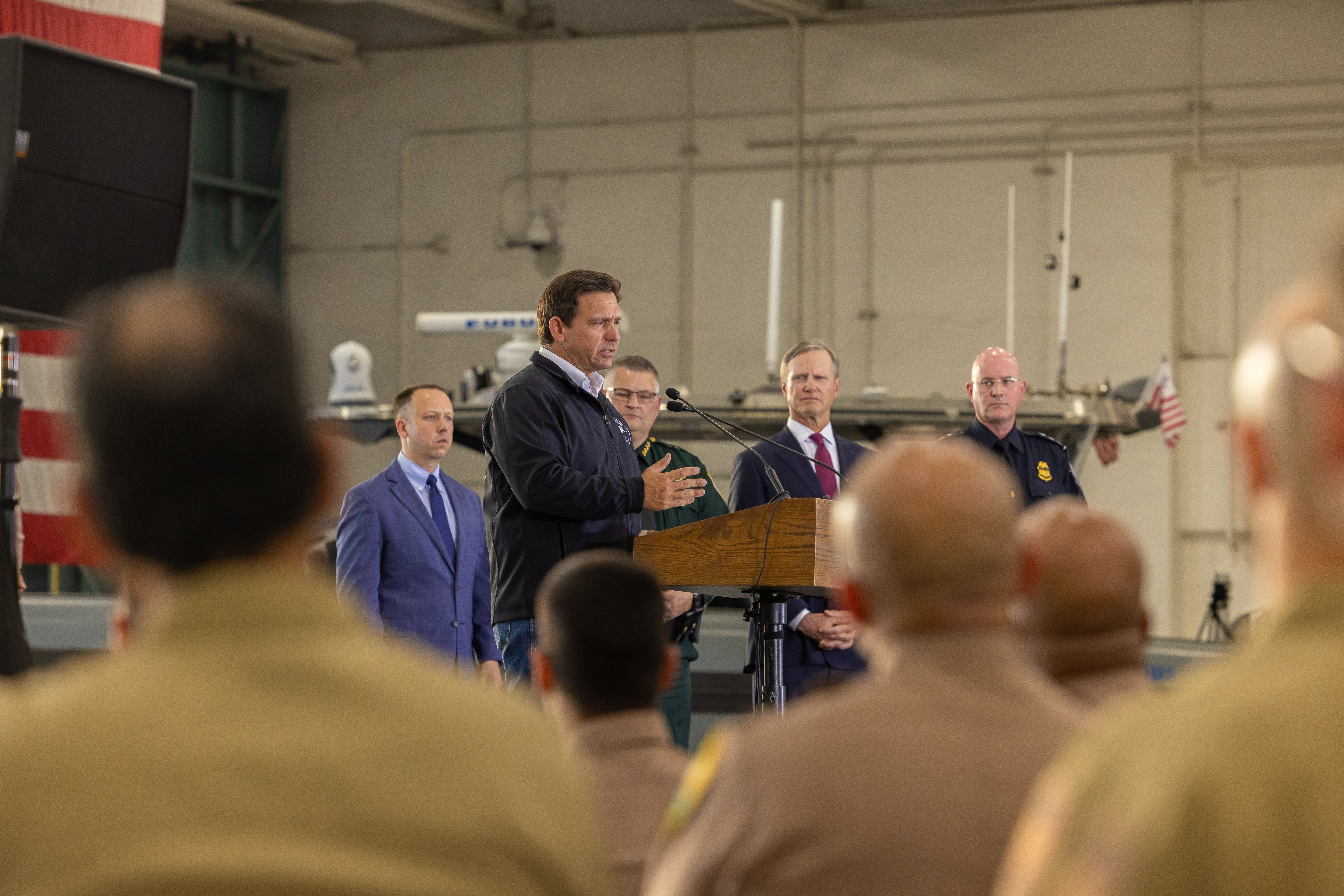 Man at a podium inside a hangar, surrounded by officials in suits and uniforms.