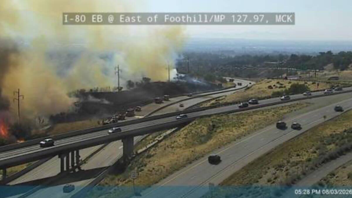 Traffic camera view of large flames and thick smoke rising from a hillside next to a busy freeway.