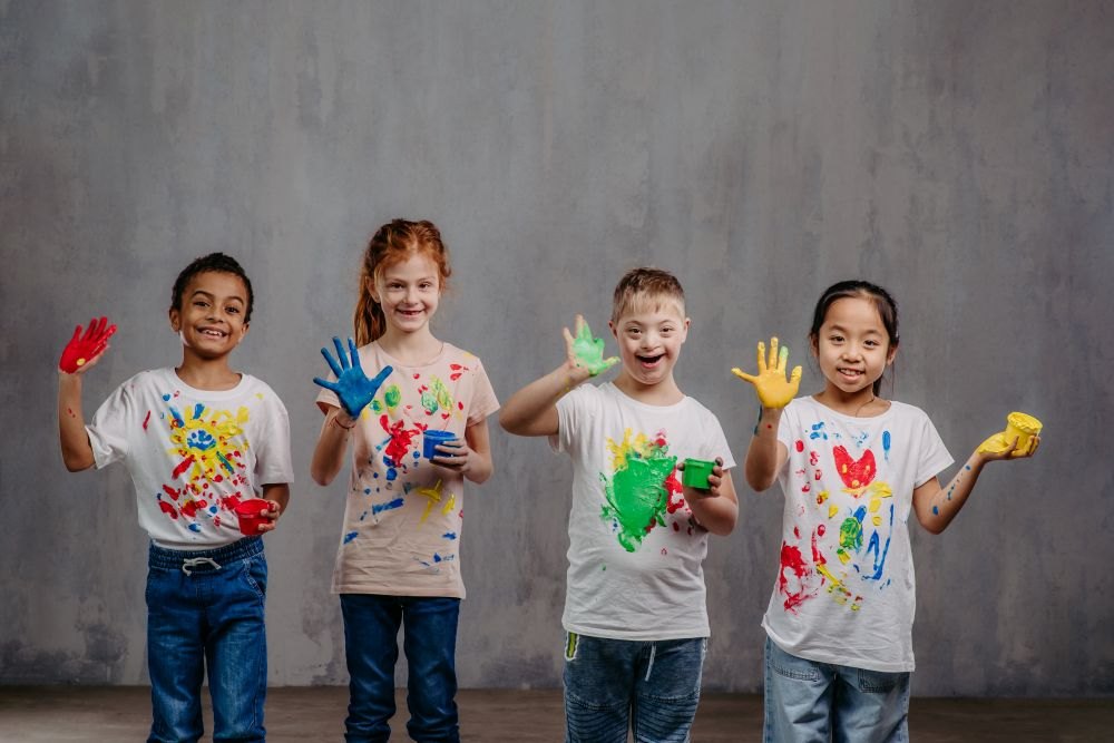 Four smiling children in paint-splattered white shirts hold up colorful painted hands.