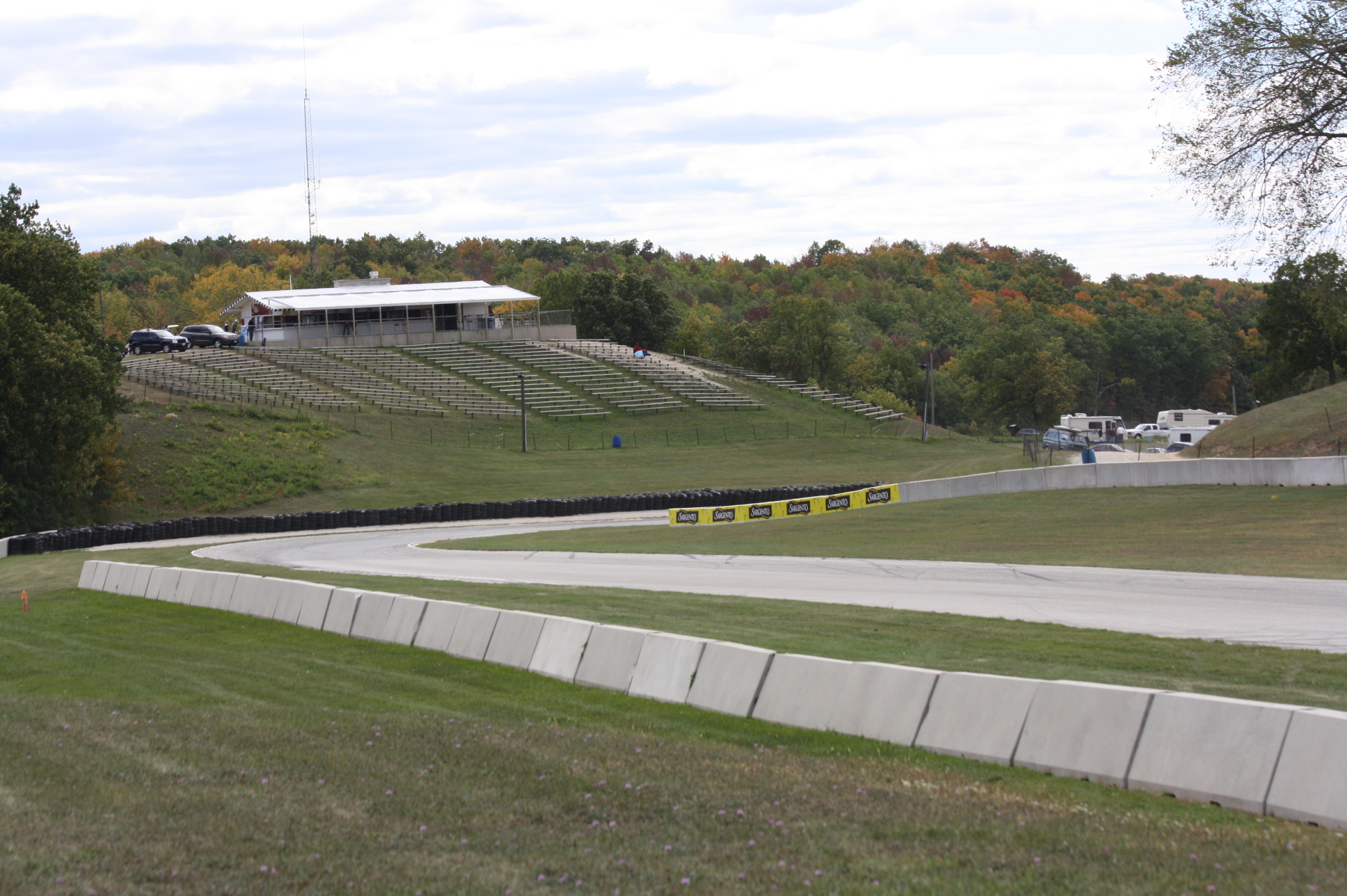 Concrete racing circuit turn with tire barriers, grandstand, and tree-lined surroundings.