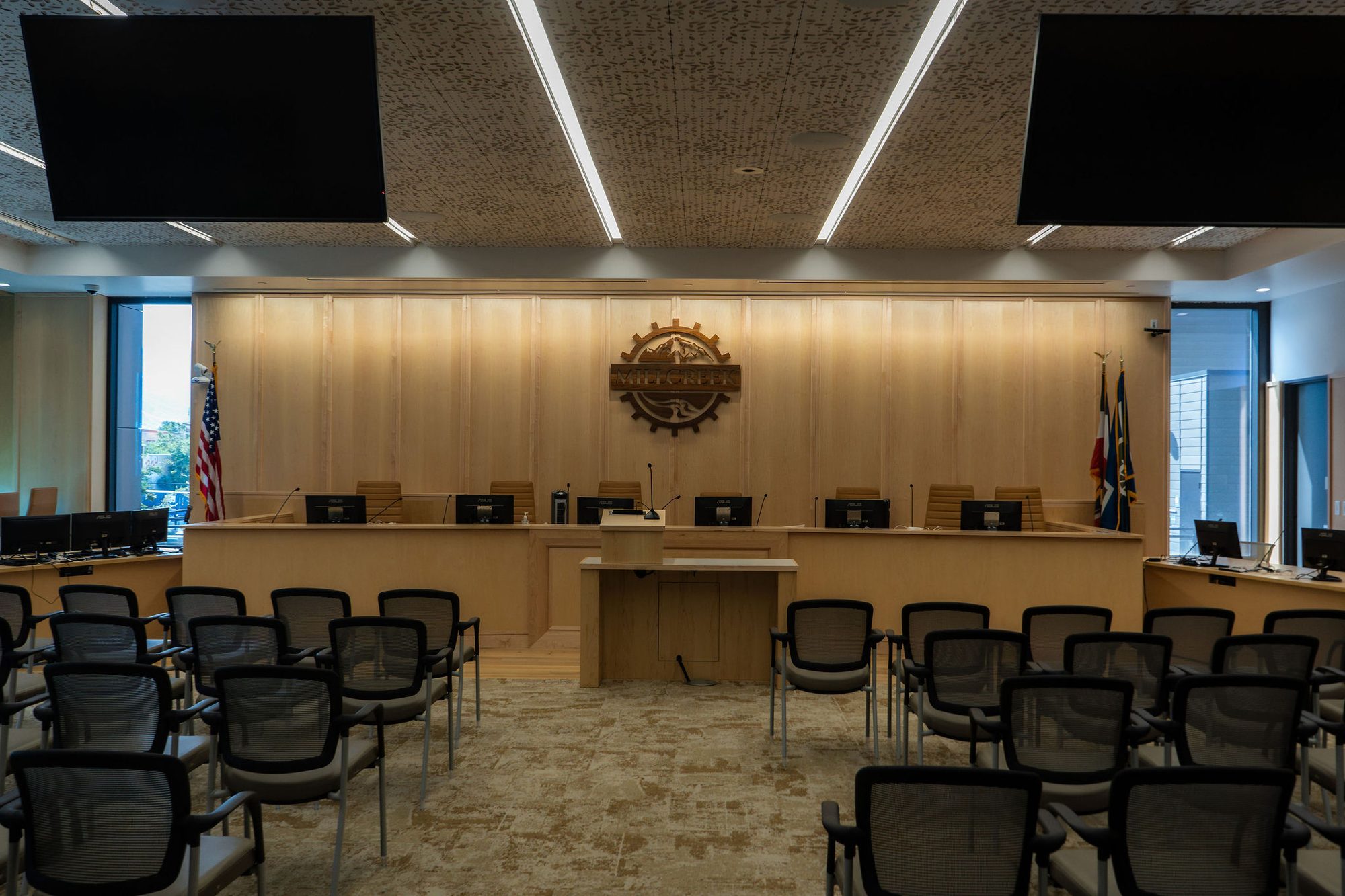 Empty city council chamber with raised dais, flags, and rows of chairs.