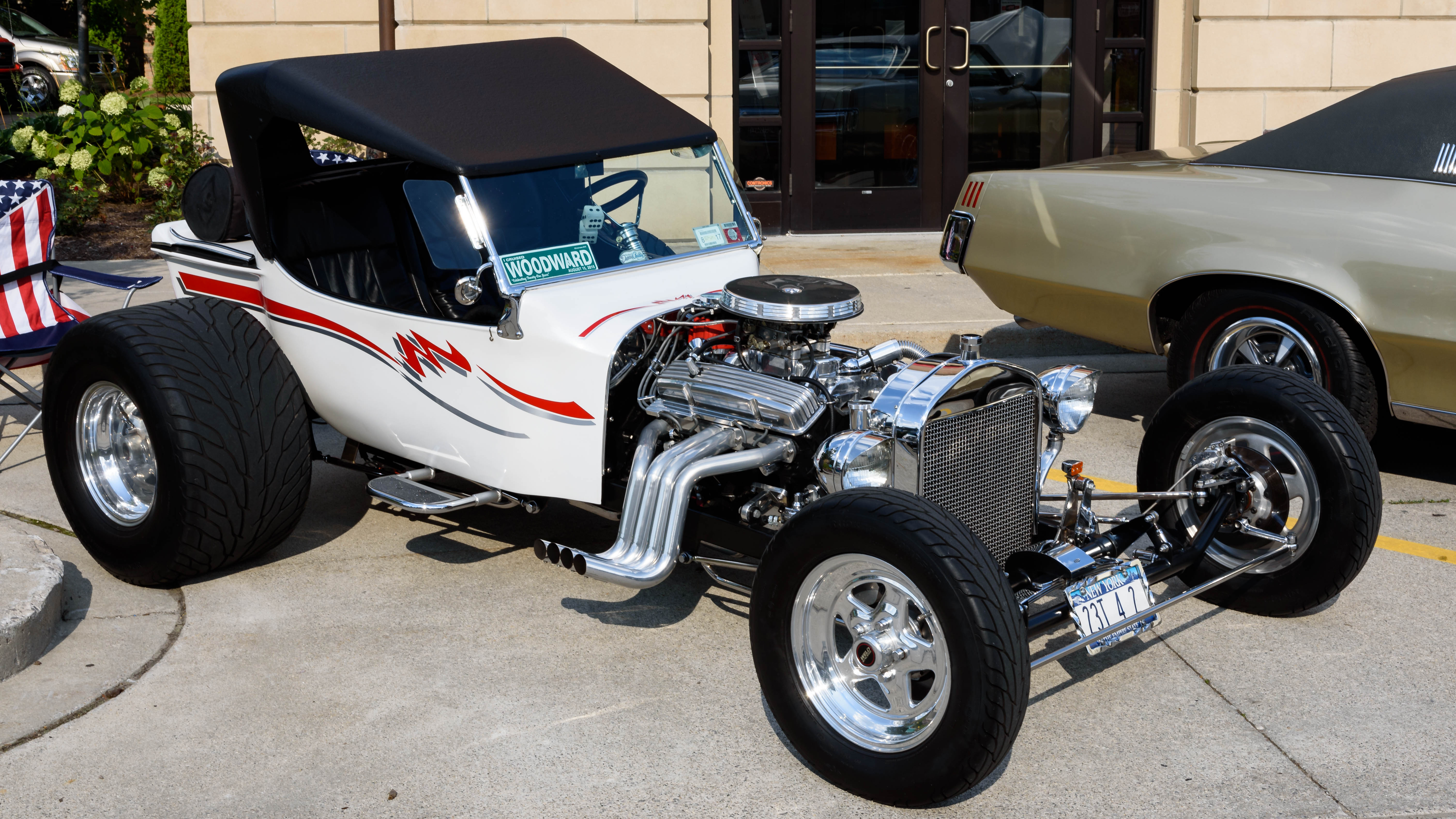 White T-bucket hot rod with exposed chrome engine and red pinstriping at an outdoor car show.