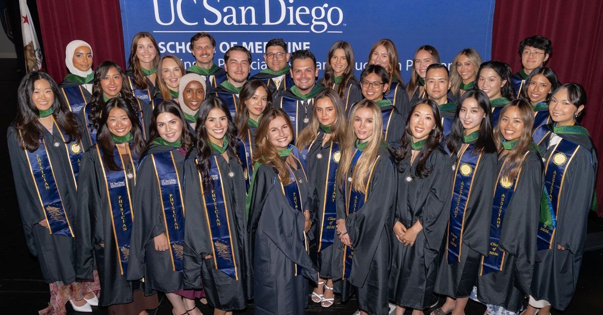 About 30 graduates in dark gowns and blue-and-gold stoles pose in front of a School of Medicine banner.