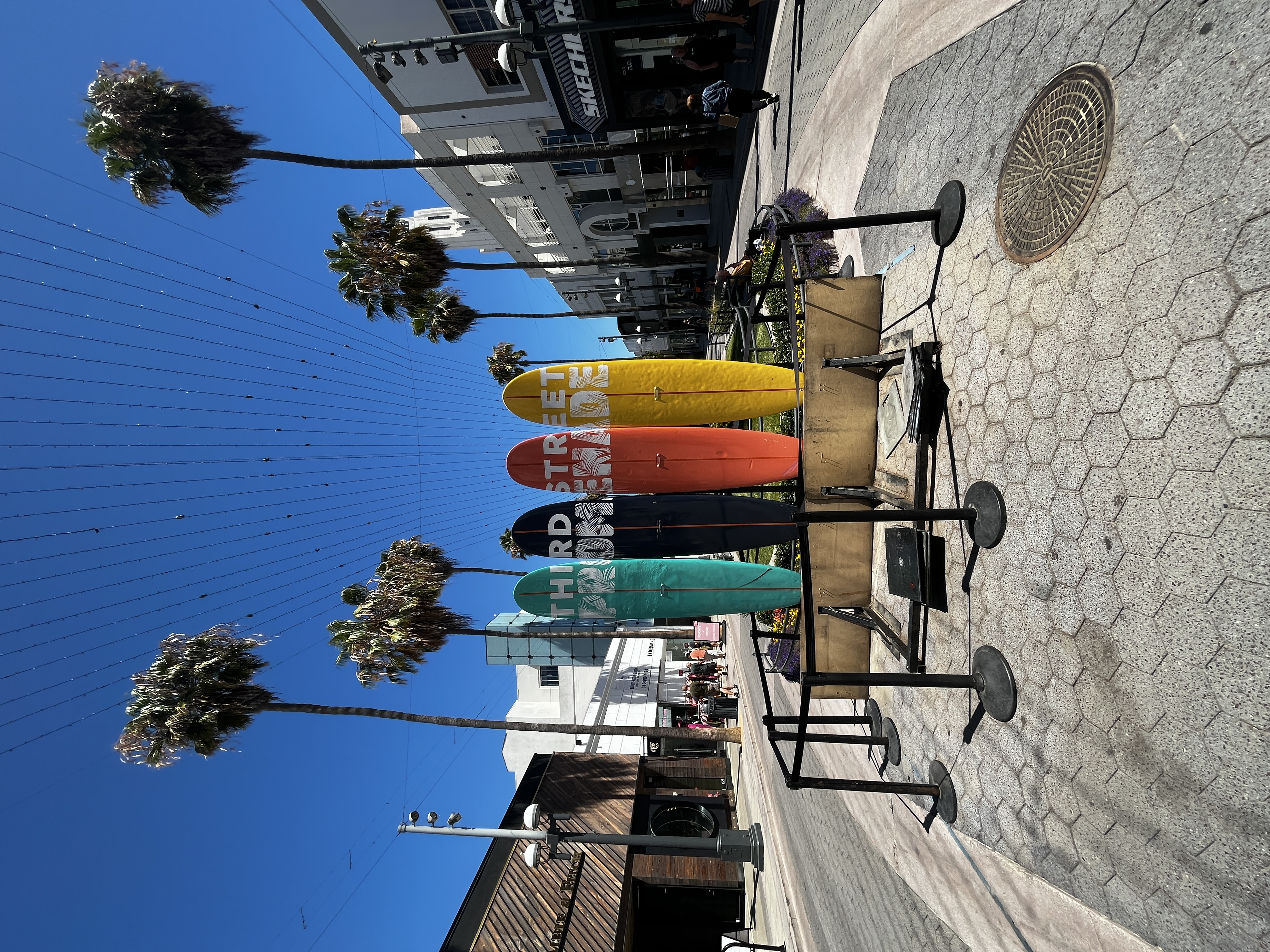 Pedestrian promenade lined with colorful surfboards, palm trees, and retail storefronts.