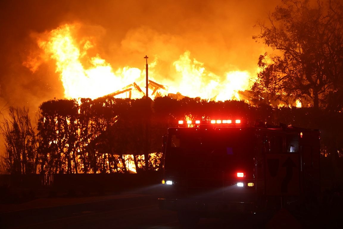 Fire truck with emergency lights parked before a large building engulfed in flames at night.