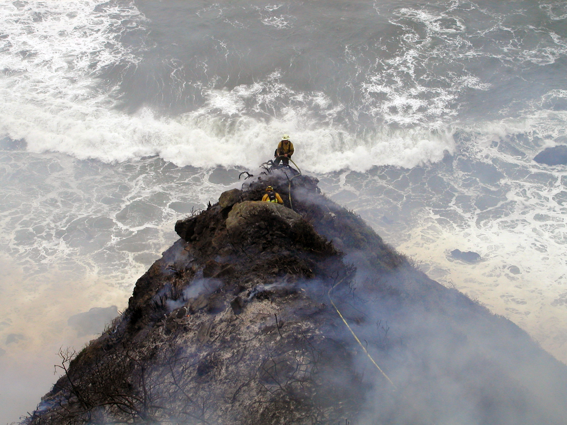 Two firefighters on a smoke-shrouded rocky cliff above the Pacific Ocean.