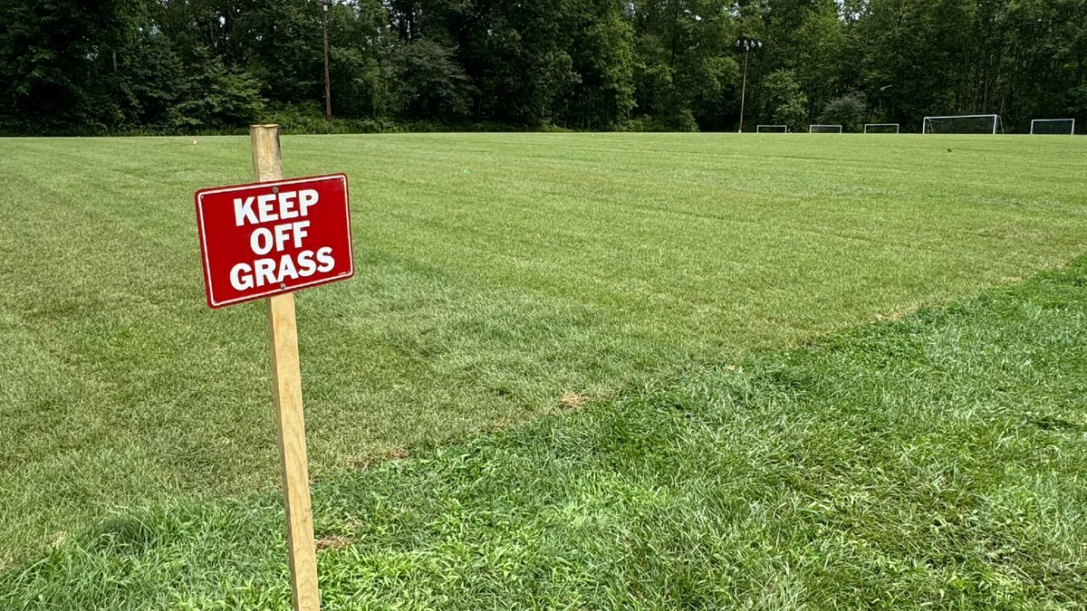 Red 'Keep Off Grass' sign on a stake in front of a freshly sodded soccer field with goals visible.