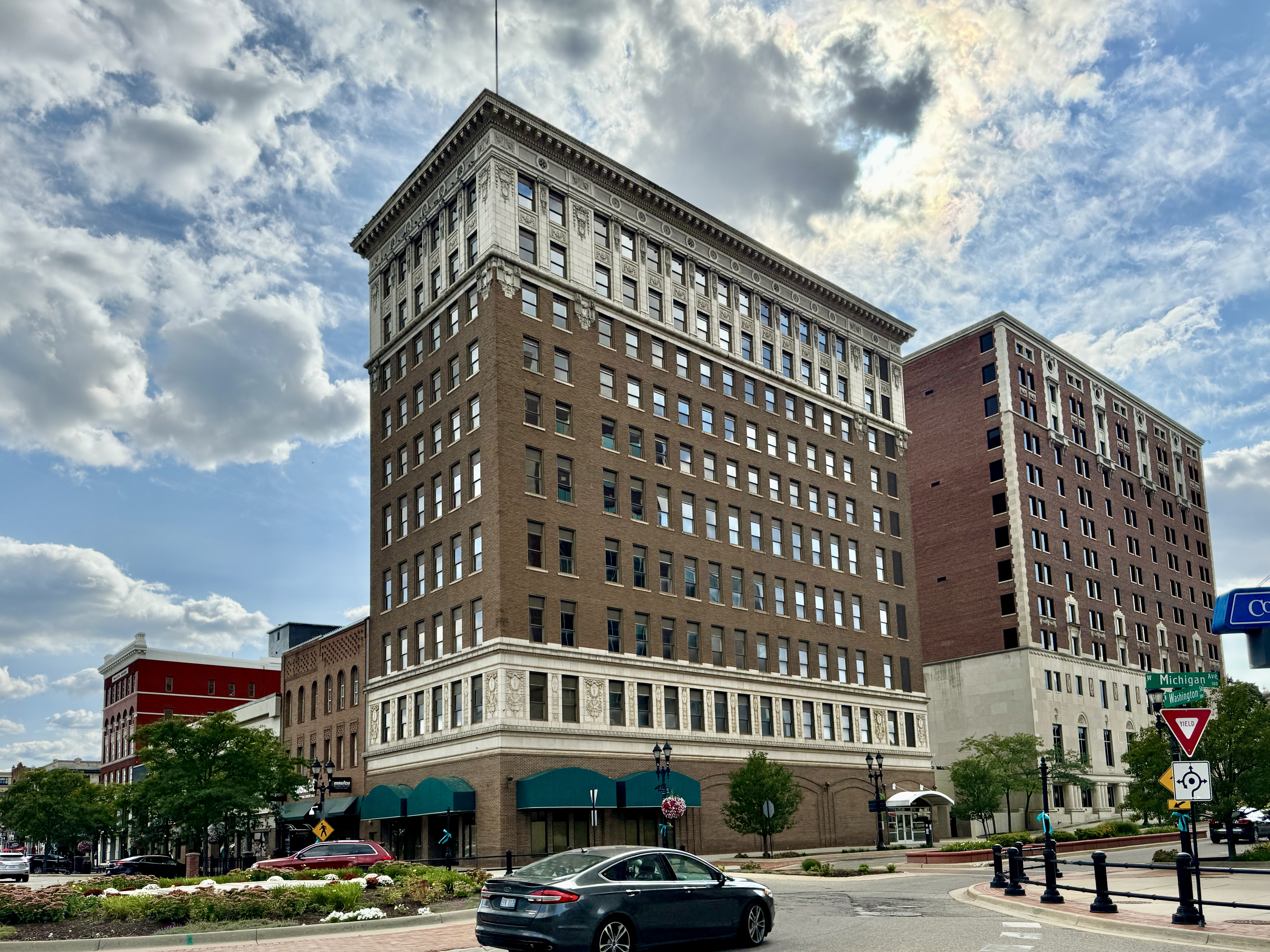 Multi-story red brick government office building on a downtown Lansing street corner with parked cars nearby.