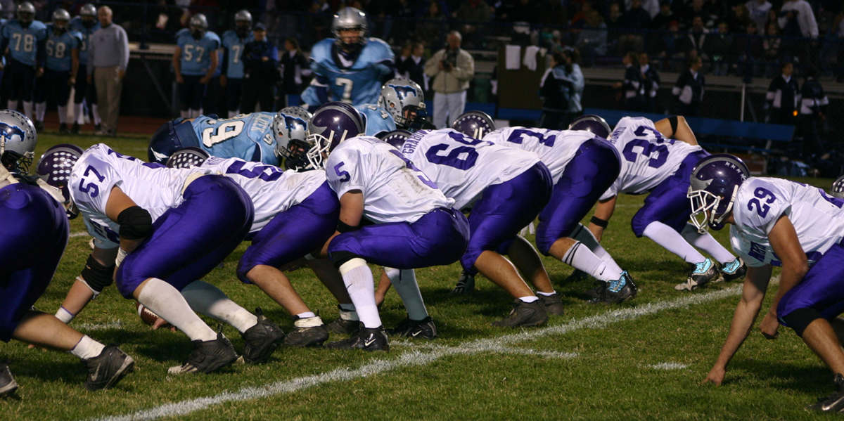 Football players in purple and white uniforms lined up at the line of scrimmage under stadium lights.