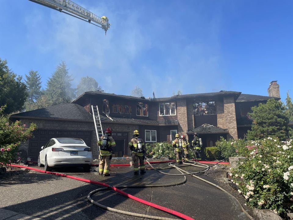 Firefighters in turnout gear work hose lines in front of a smoke-damaged two-story brick home with an aerial ladder extended above.