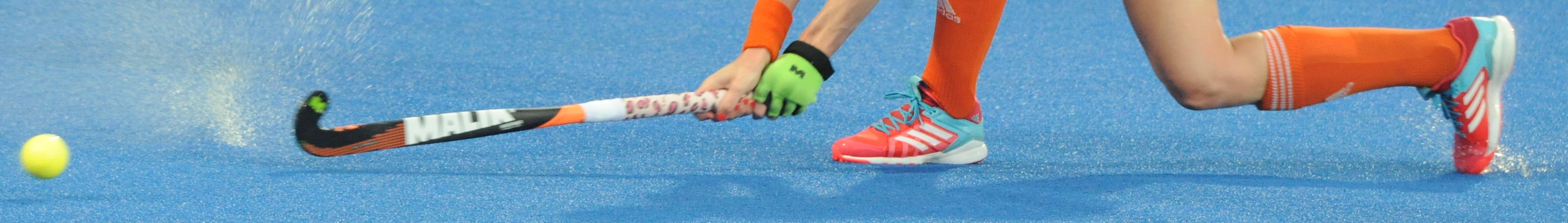 Field hockey player's legs and stick in motion on blue artificial turf near a ball.
