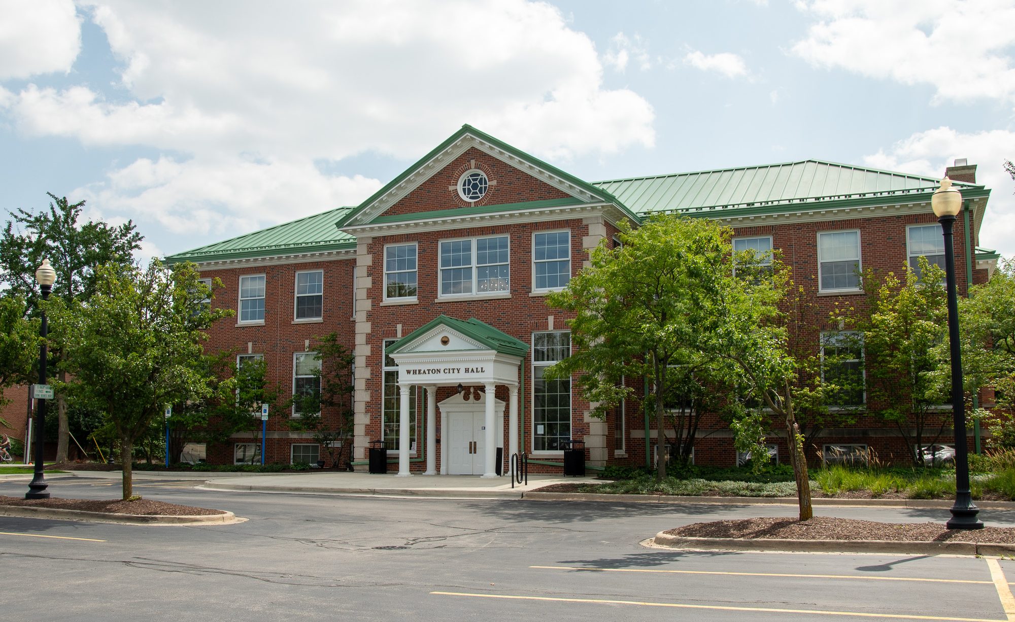 Two-story red brick city hall building with green roof at a street corner.
