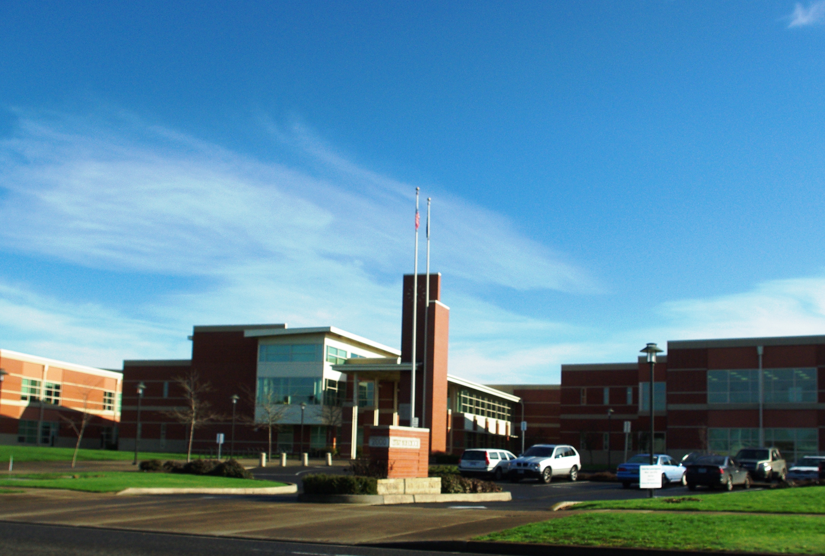Red brick high school building with two flagpoles and a parking lot under blue sky.