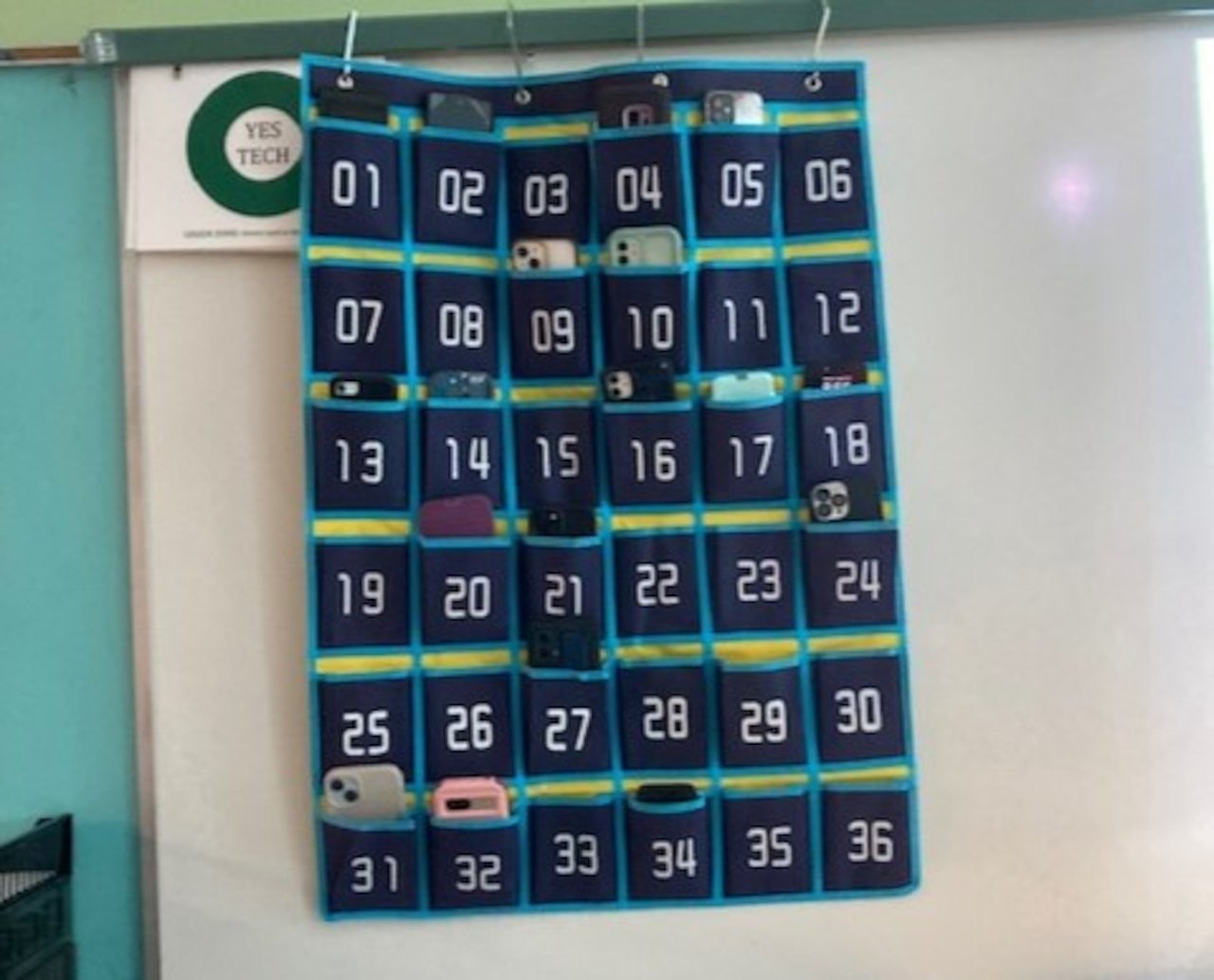 Numbered pocket chart on a whiteboard holding dozens of student smartphones.