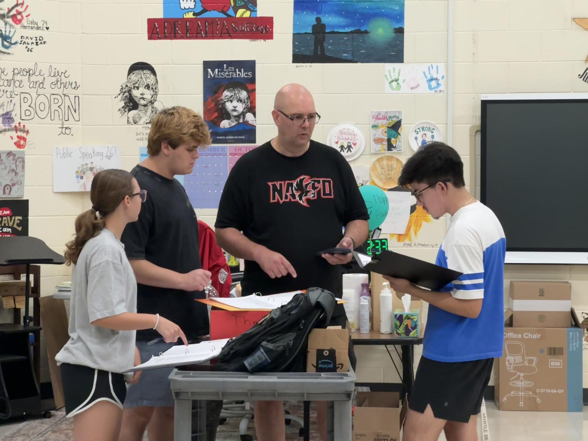 An adult in a black shirt reviews materials at a table with three students in a classroom with theater posters.