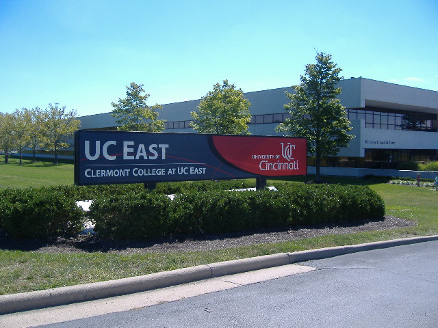 Campus sign reading UC East Clermont College University of Cincinnati, with building and trees behind.