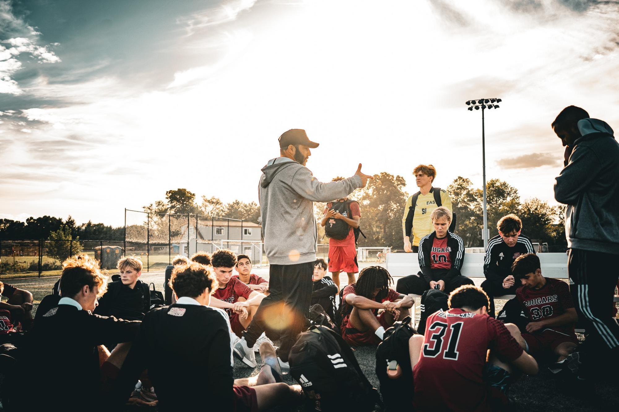 Coach addresses seated male soccer players in a circle on an outdoor field at dusk.