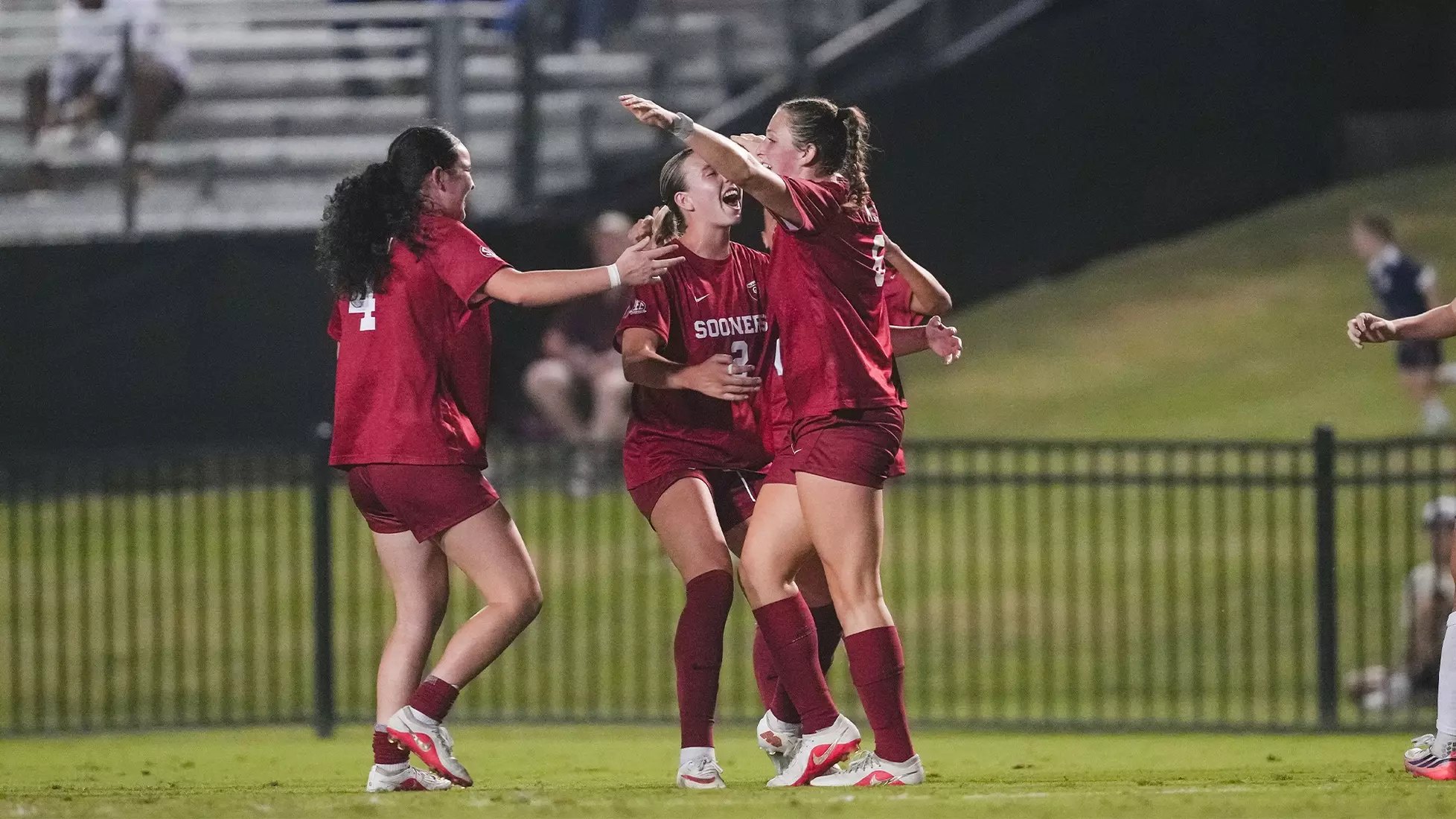 Three soccer players in red uniforms embrace and celebrate on a grass field at night.