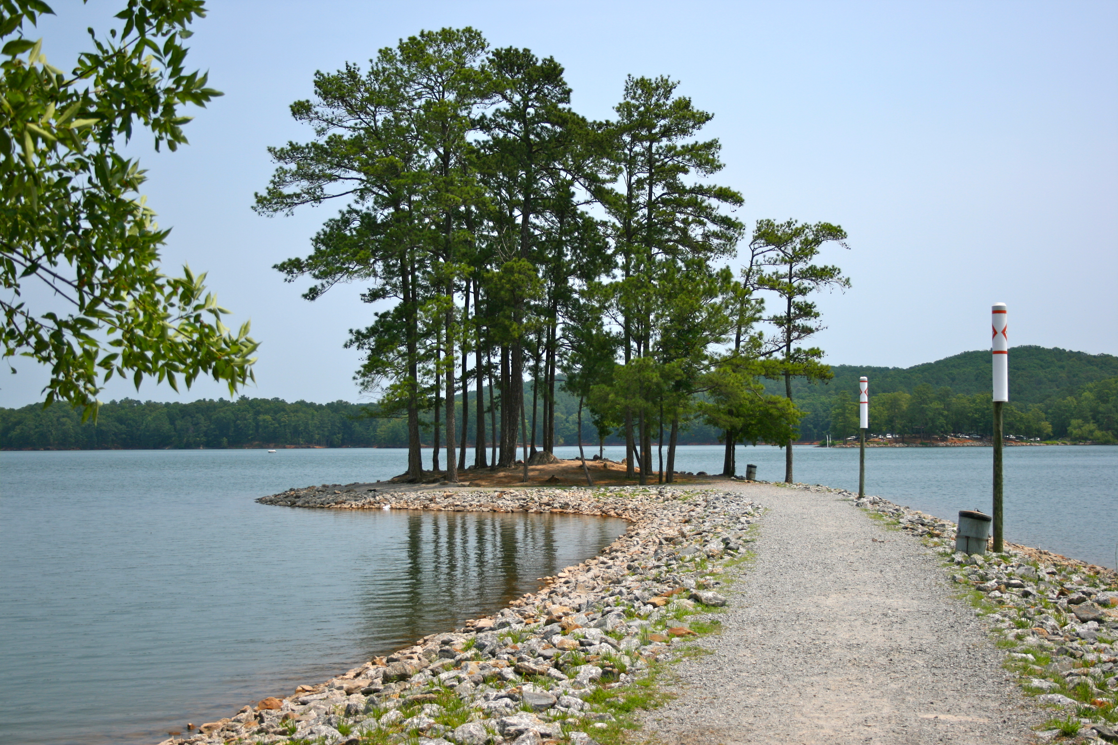 A rocky causeway extends into a calm lake lined with tall pine trees and a navigational marker post.