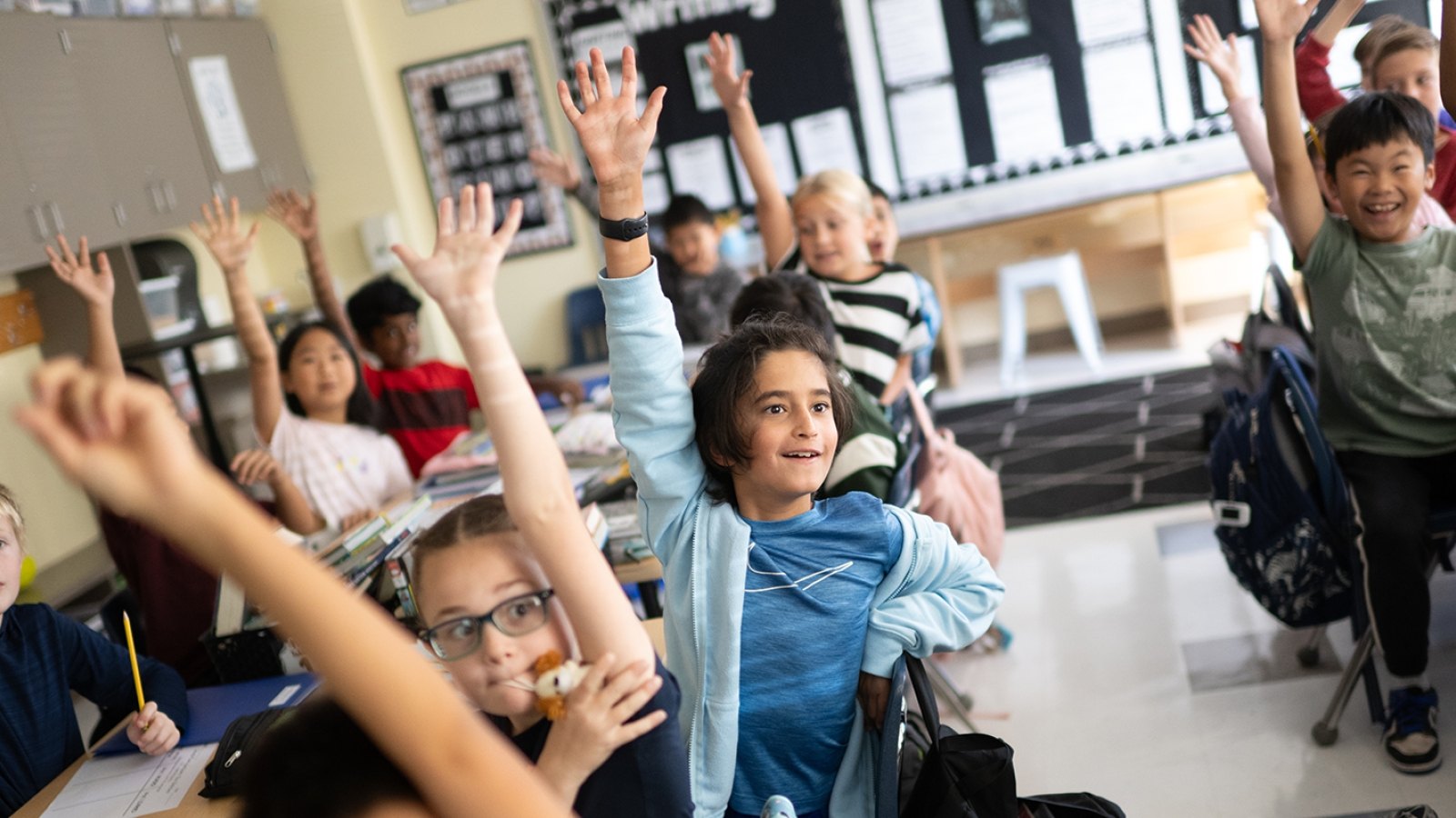 Elementary school students sitting at desks with hands raised in a bright classroom.