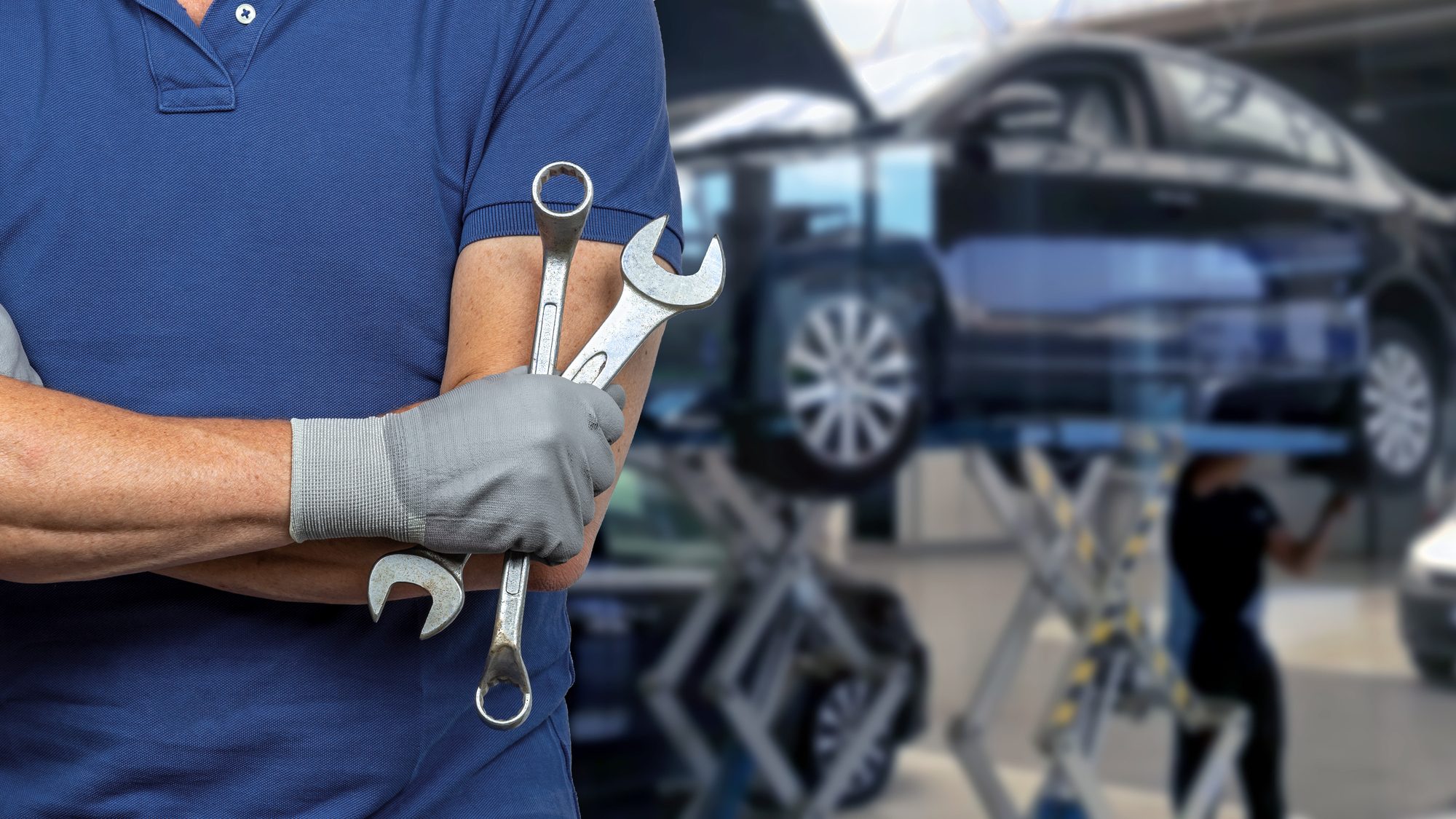 Mechanic with crossed arms and holding wrenches in auto repair workshop.