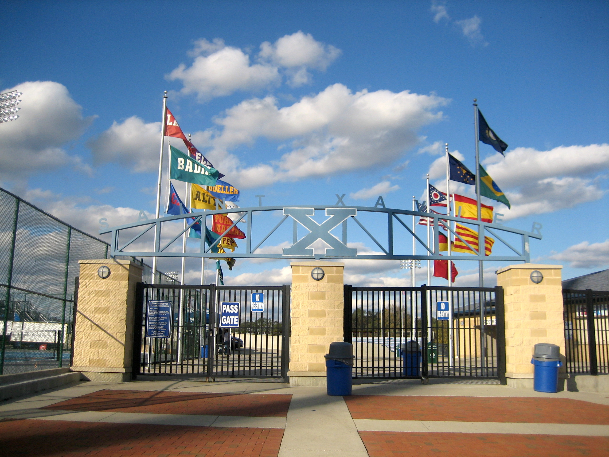 A stone and metal entrance gate to a football stadium with an X logo arch and flags flying above.